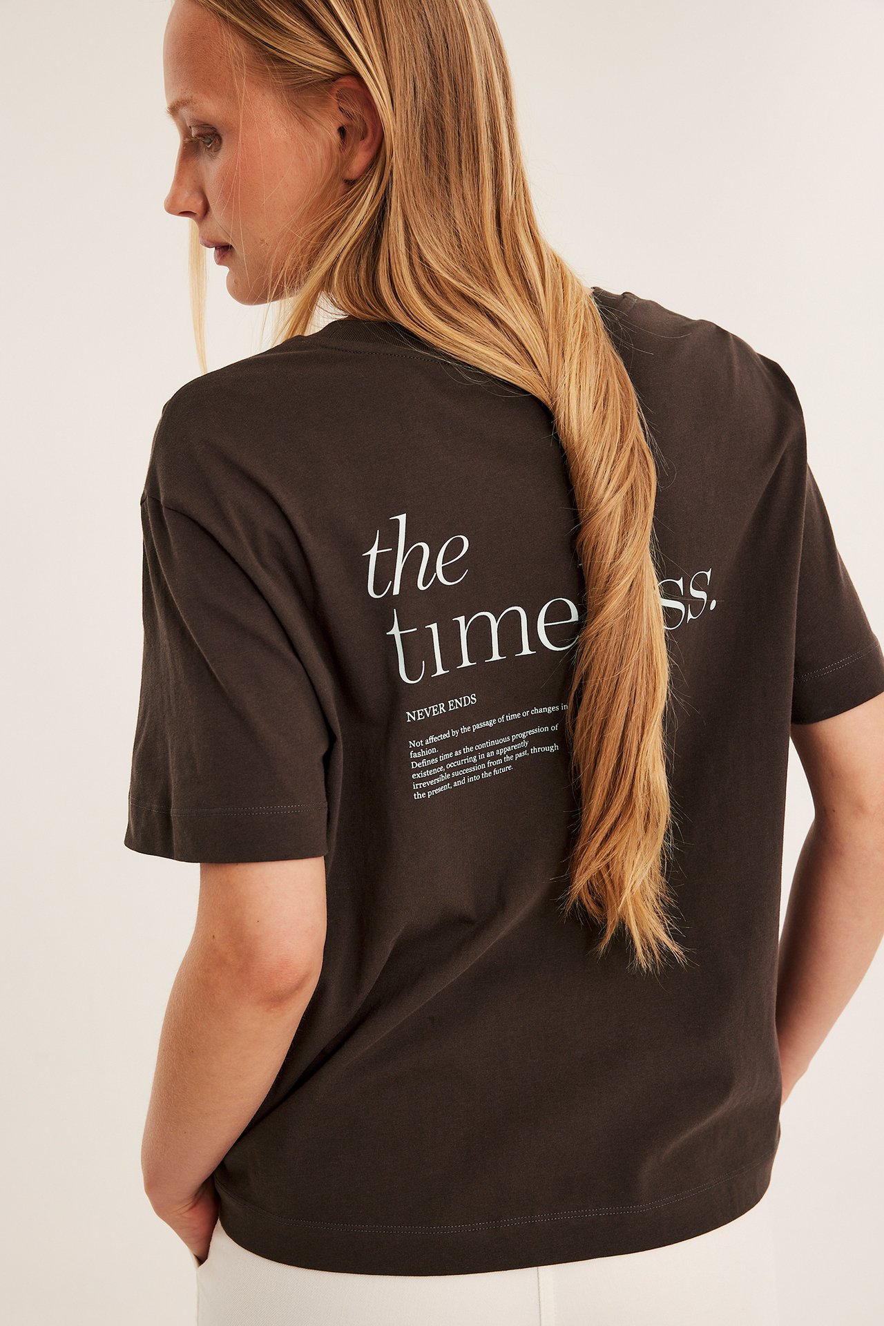Nainen yllään tummanruskea puuvillainen t-paita 'the timeless.' -tekstillä painettuna takana, taka-/sivukuva.