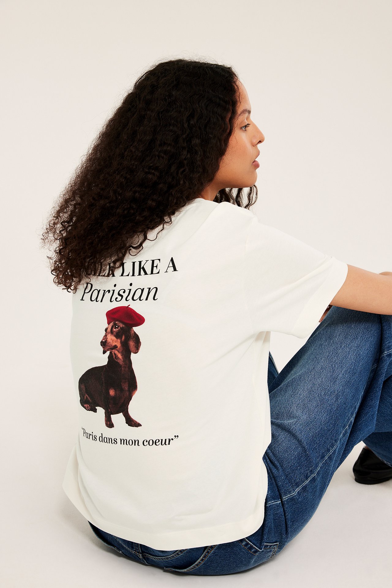 Bakifrån: Kvinna i vit bomulls-t-shirt med tryck "DREAM LIKE A Parisian", hund, och blå jeans.
