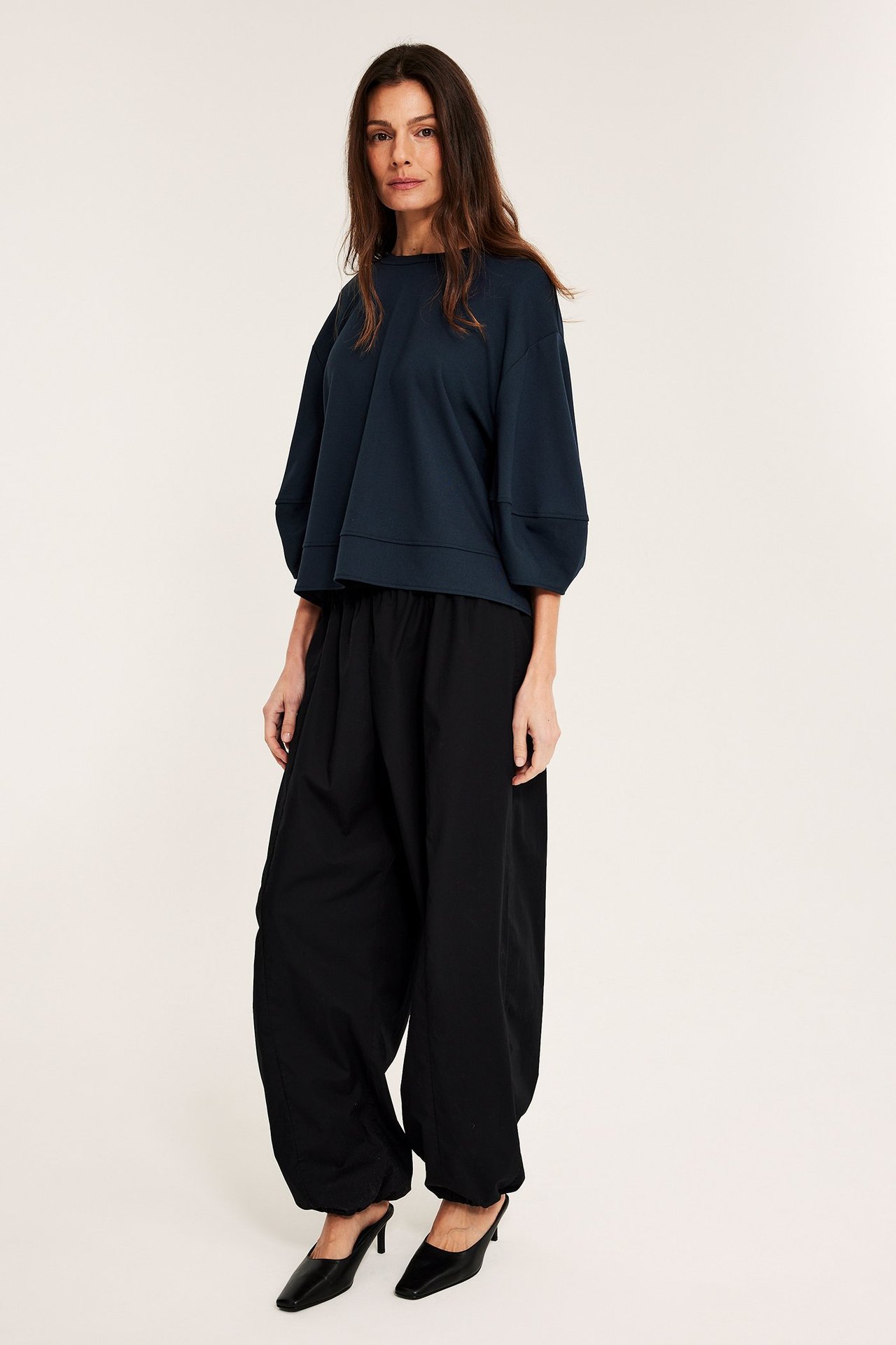 Widok z przodu: Kobieta w granatowym luźnym topie punto z bufiastymi rękawami, czarnych szerokich spodniach i czarnych klapkach.