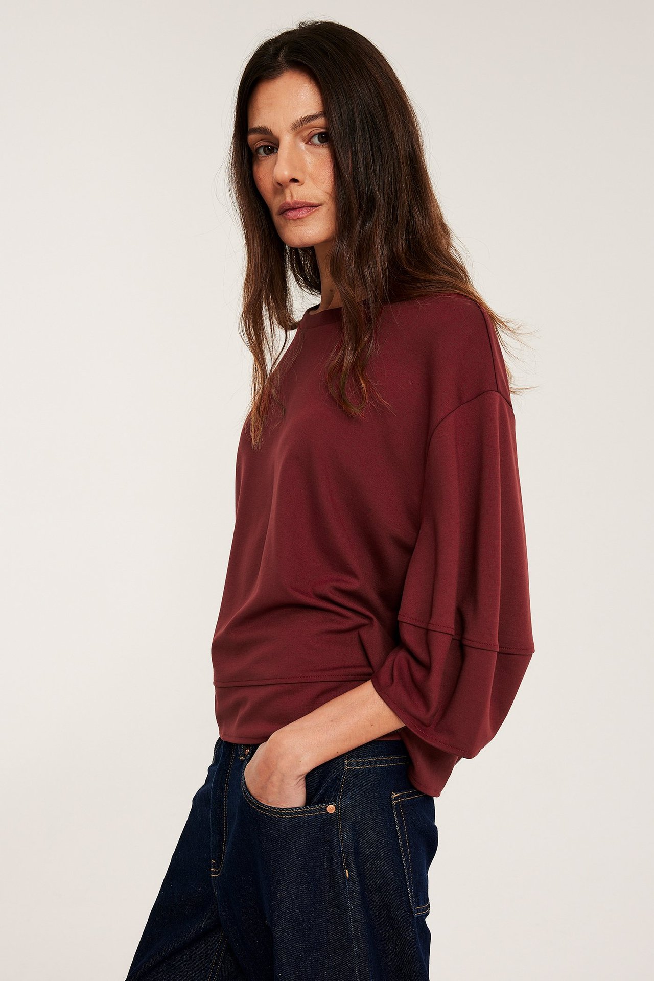 Edestä, nainen yllään löysä punainen punto-toppi pöyhkeillä hihoilla ja tummansiniset farkut, käsi taskussa.