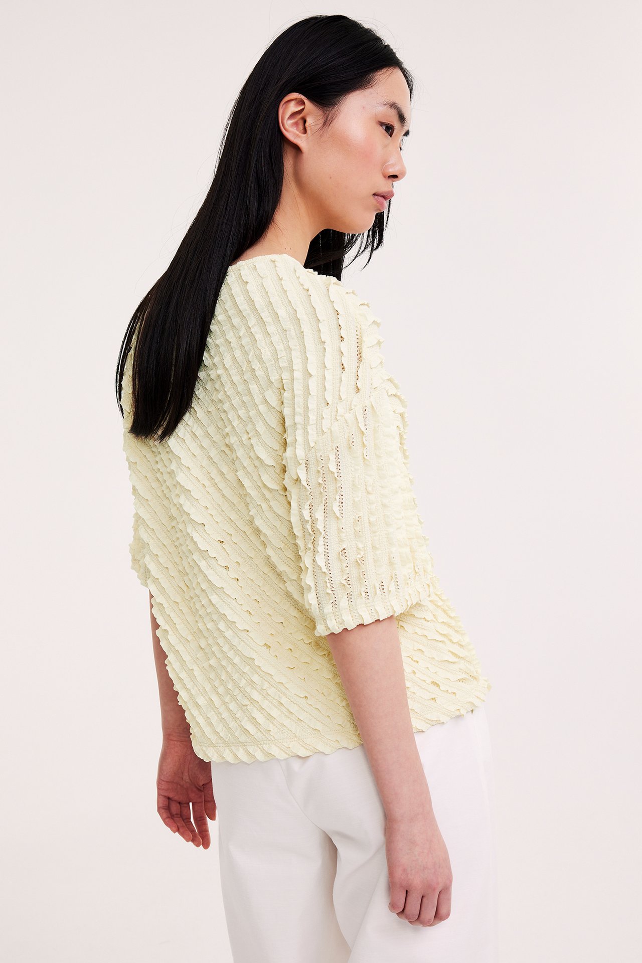 Sivultapäin nainen yllään vaaleankeltainen strukturoitu löysä lyhythihainen toppi ja valkoiset housut.