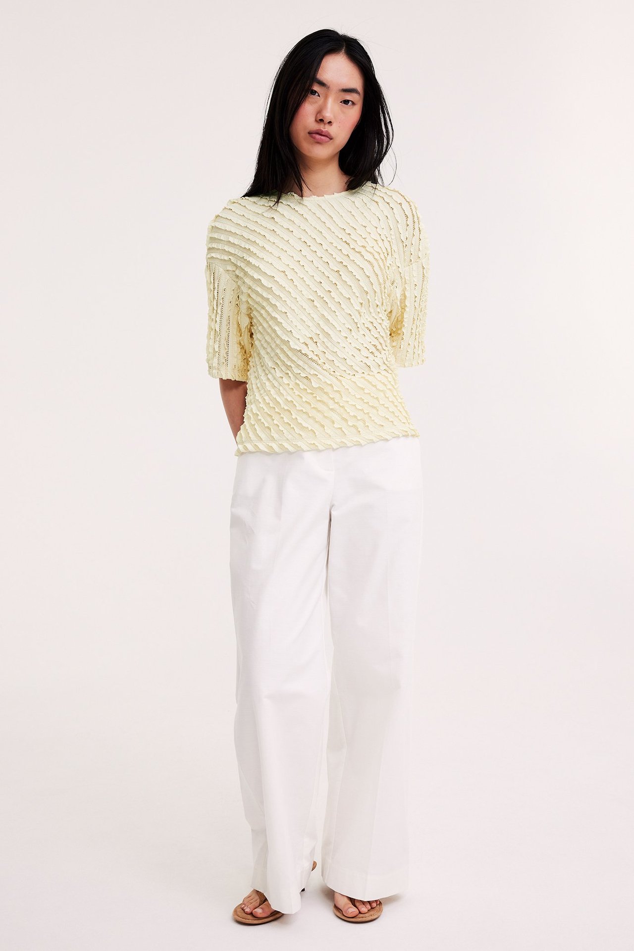 Widok z przodu: kobieta w jasnożółtym strukturalnym topie, białych szerokich spodniach i brązowych sandałach.