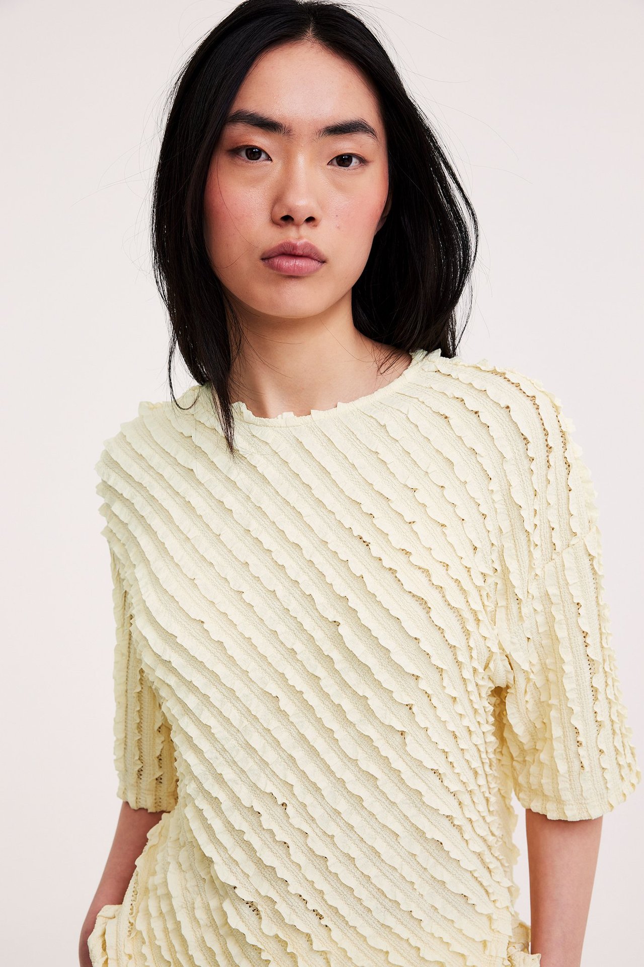 Zbliżenie z przodu na kobietę w jasnożółtym, luźnym, krótkim topie z diagonalnymi strukturalnymi falbankami.