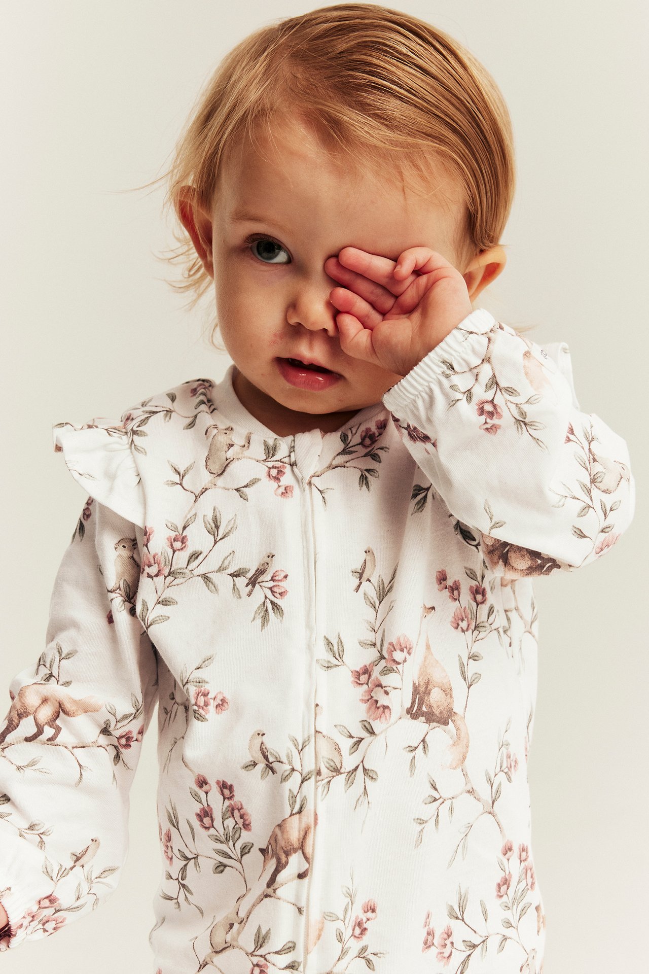 Frontvy, baby i vit långärmad pyjamas med rosa blommigt, djurmönster och volangkrage.