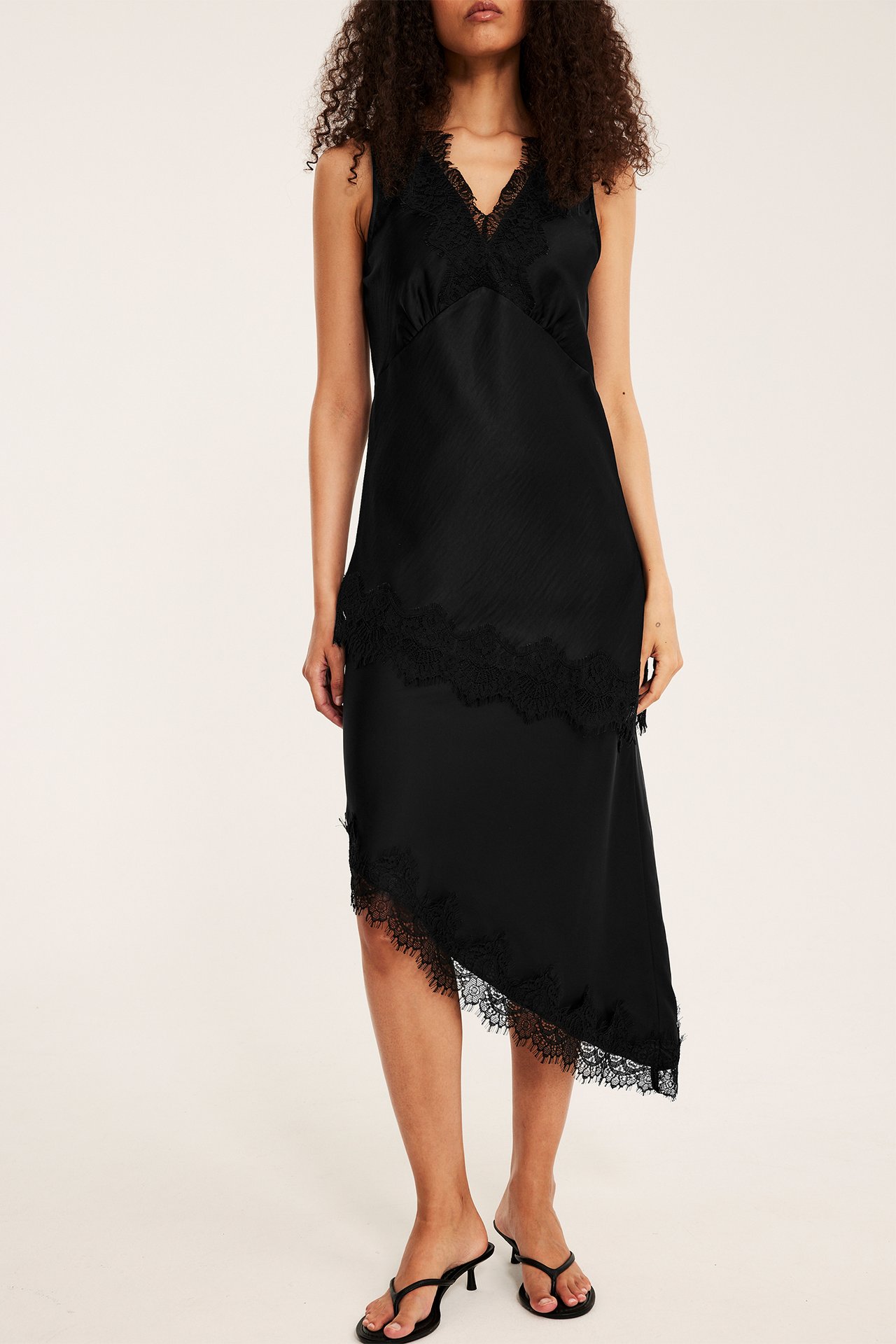 Framsida av en kvinna i svart satinklänning med spets och asymmetrisk fåll, med svarta klackar.
