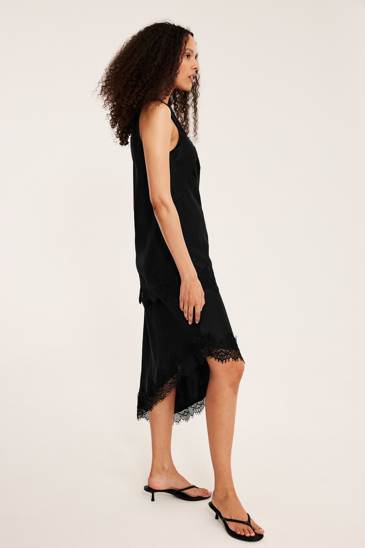 Sidovy av kvinna i svart satinklänning med asymmetrisk spetskant, bär svarta sandaler.