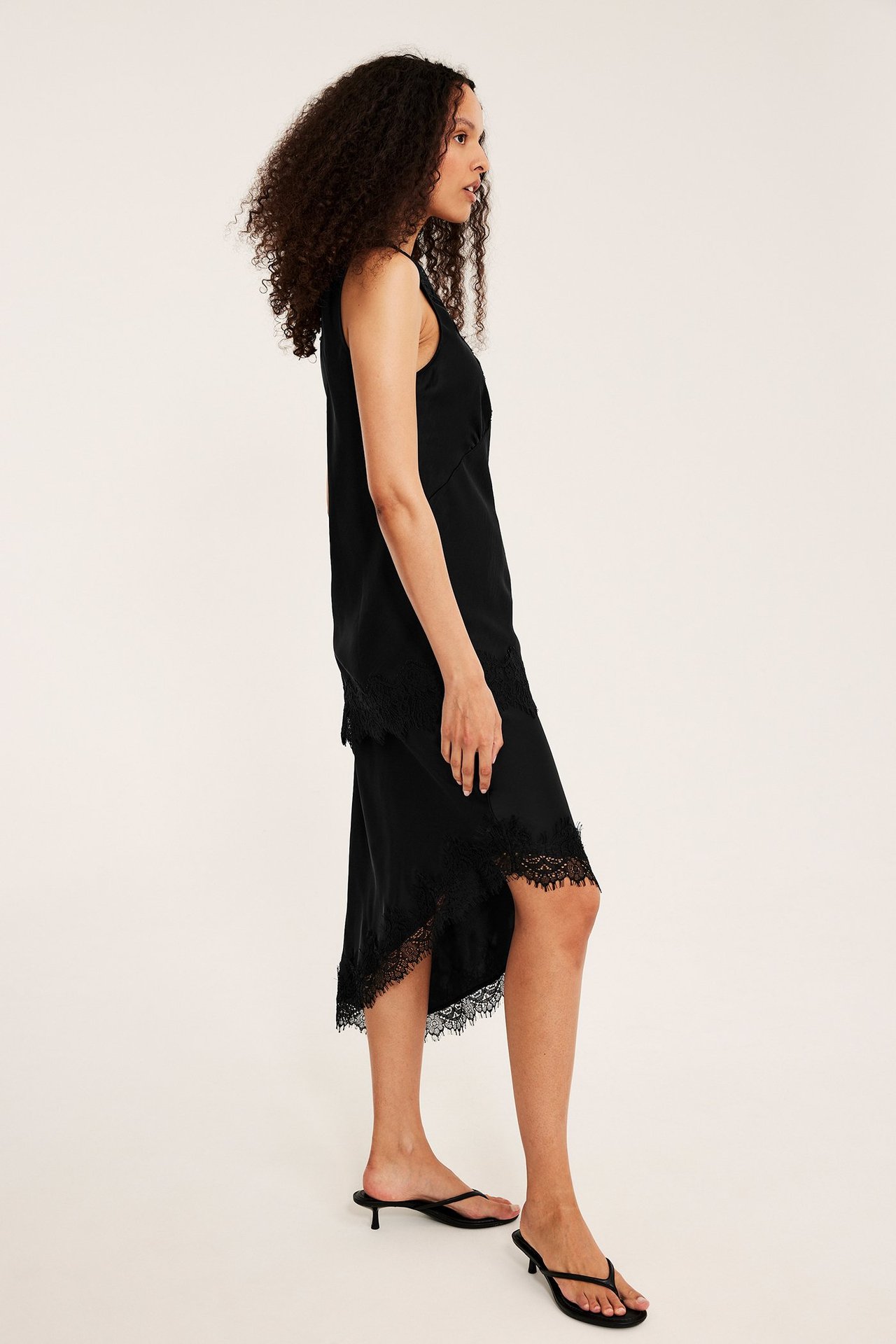 Widok z boku kobiety w czarnej satynowej sukience z asymetryczną koronką, czarne sandały.