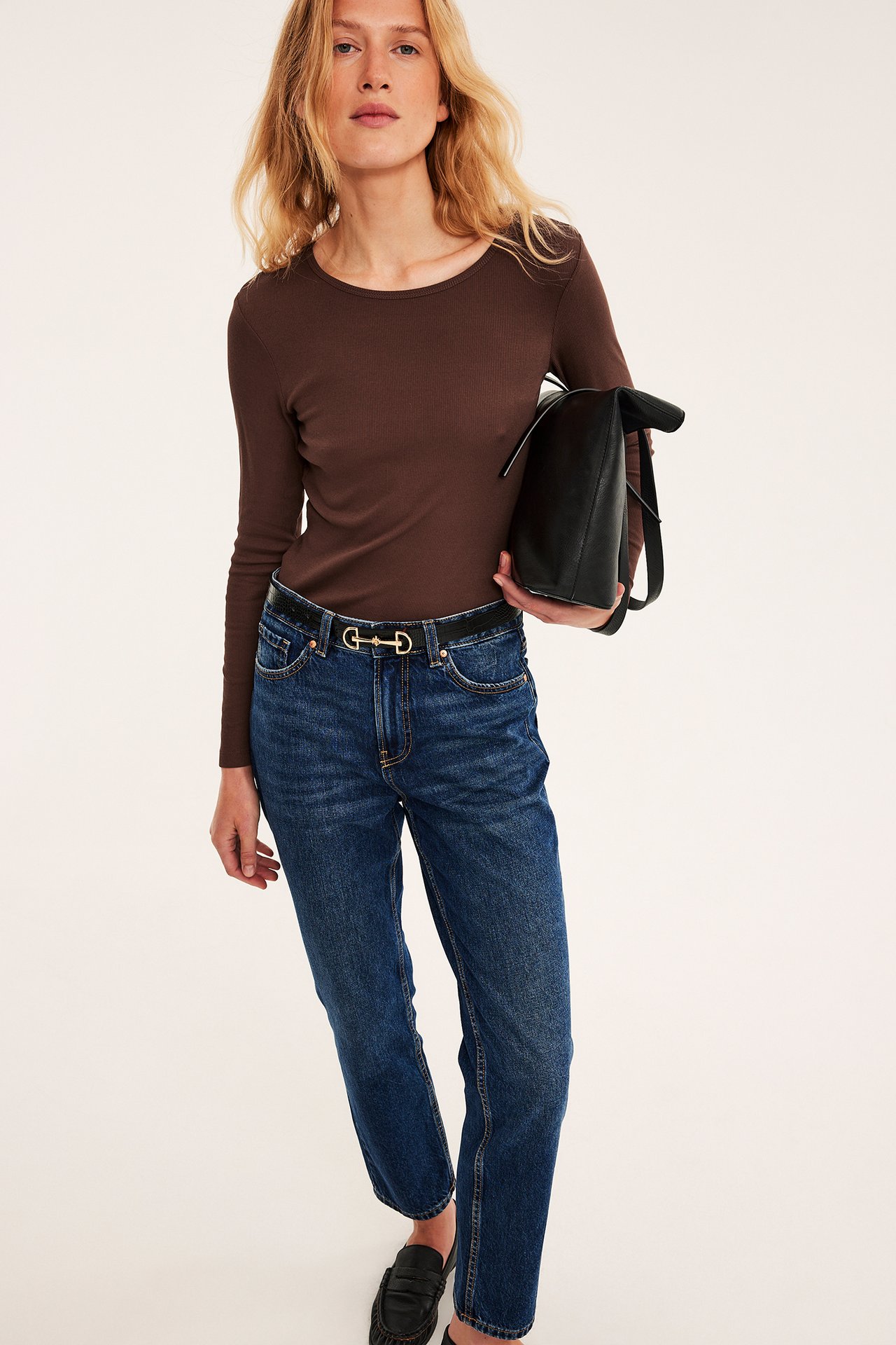 Kvinnlig modell bär brun långärmad topp, blå jeans, svart bälte, svarta loafers, svart handväska.