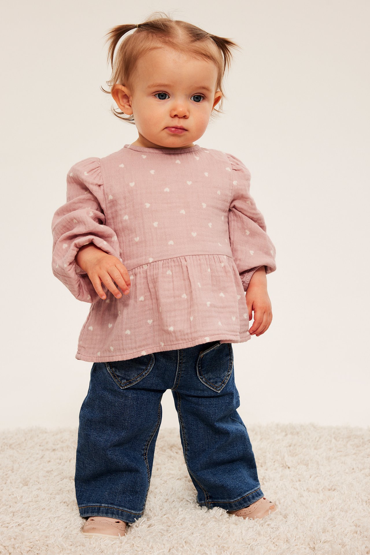 Frontbilde av baby i rosa hjertemønstret muslin peplumtopp, blå jeans og rosa sko.