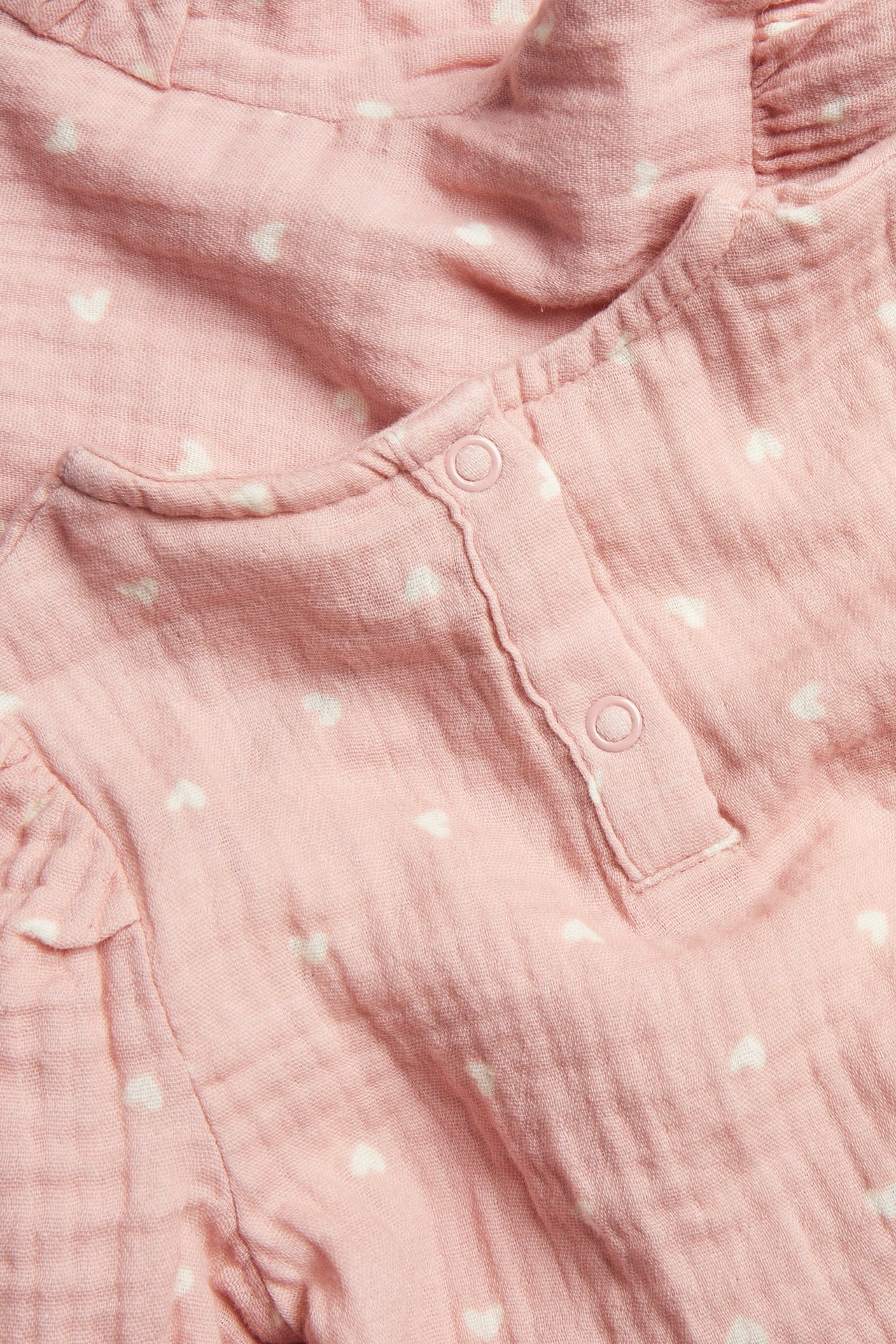 Nærbilde av rosa babyplagg med hvitt hjertemønster, krøllete stoff og trykknapper.