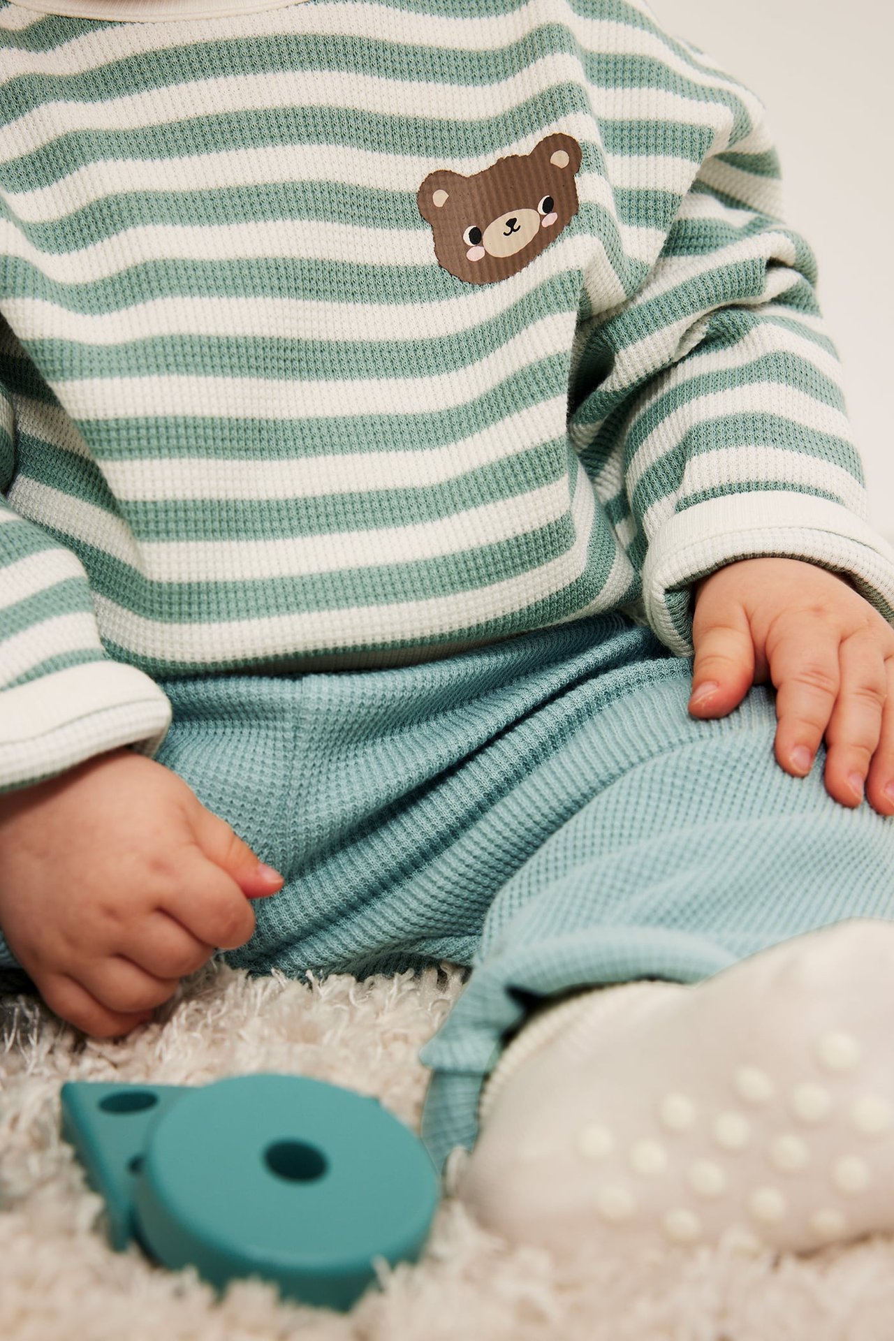 Nærbilde: Baby har på seg grønn-hvitstripet vaffelstrikket topp med bjørnelapp og turkise bukser.