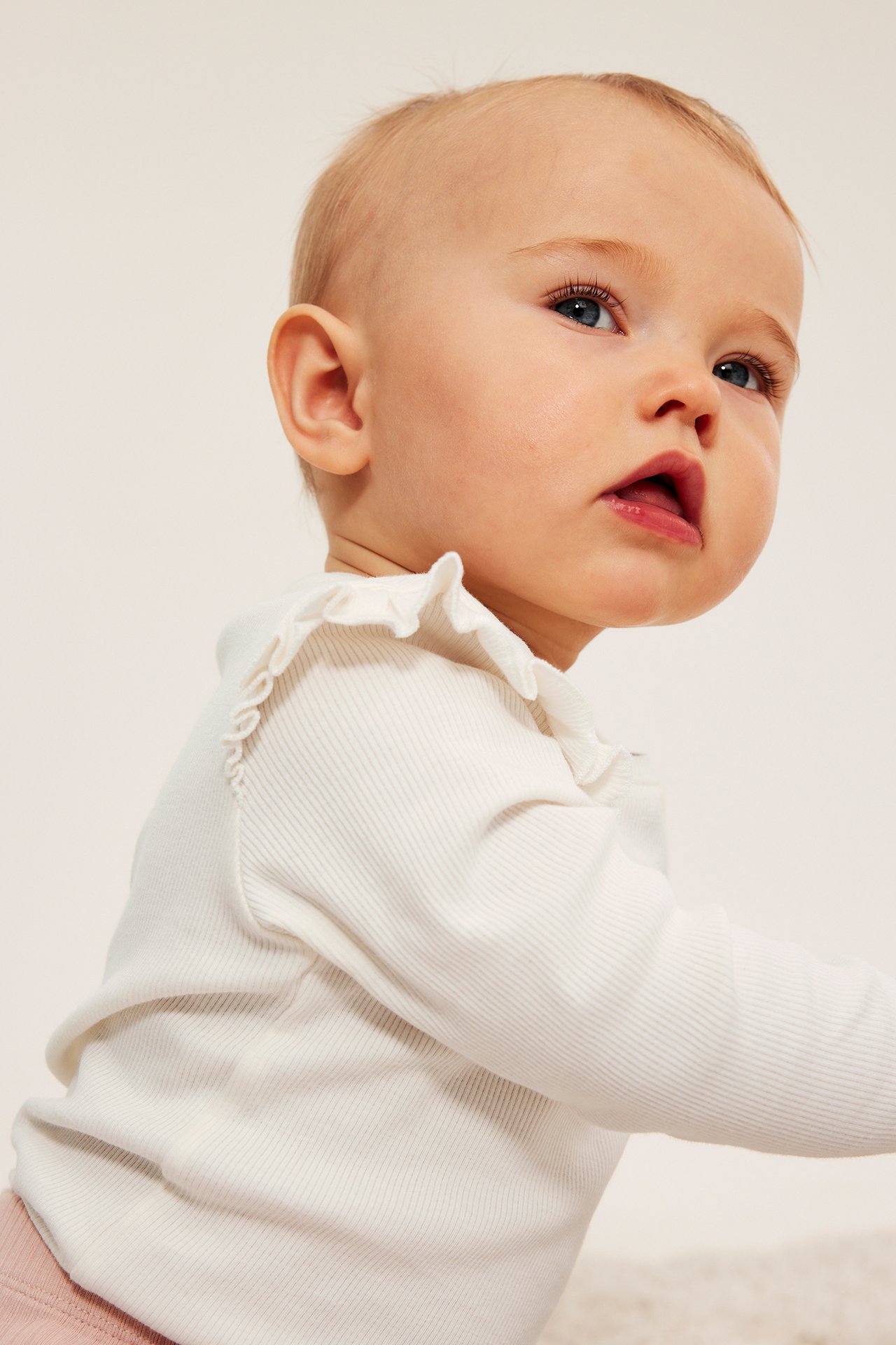 Närbild på en baby i en vit ribbad långärmad topp med volanger och rosa underdel.