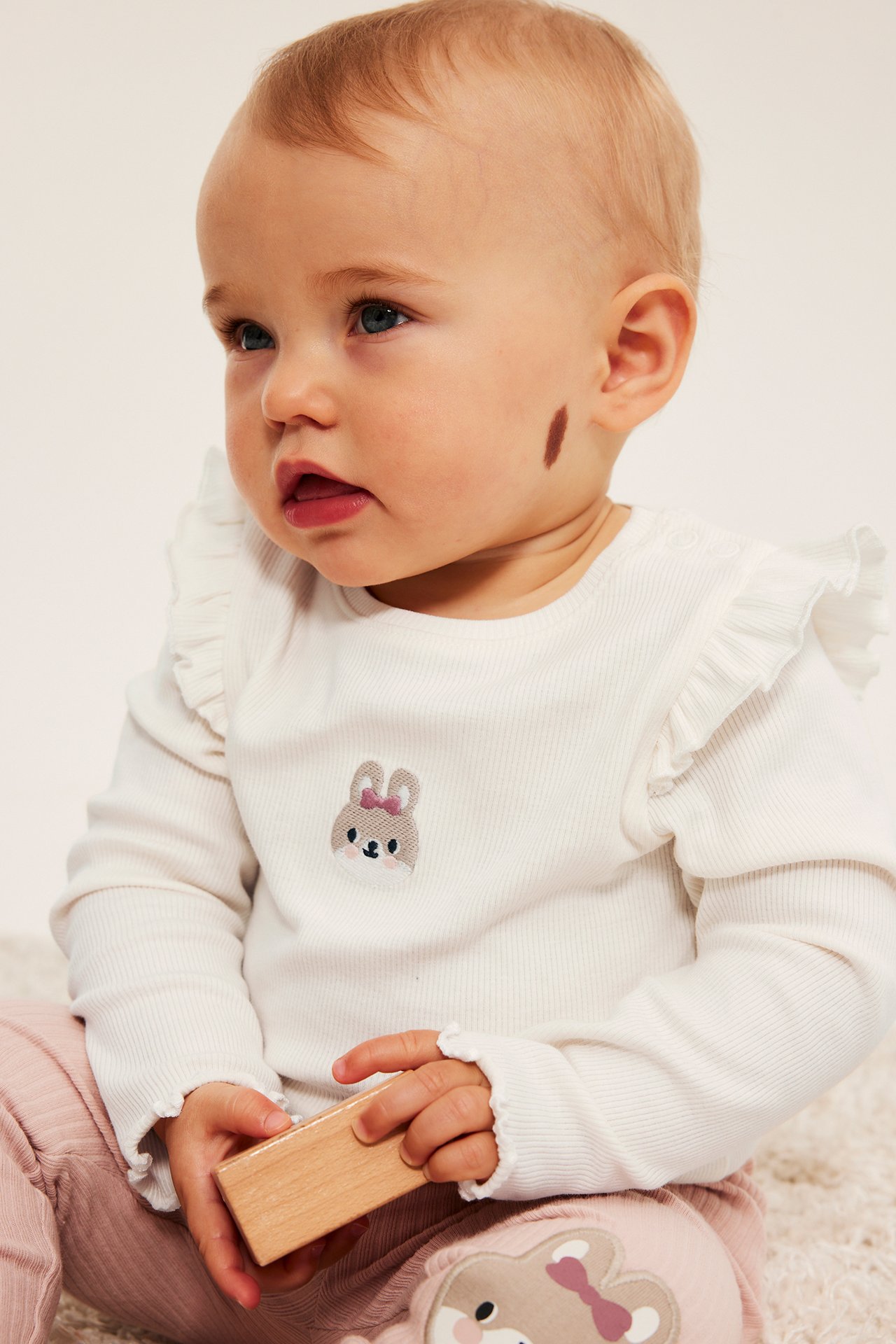 Baby bär vit ribbad långärmad topp med kanin, rosa ribbade leggings med lapp, håller träkloss.