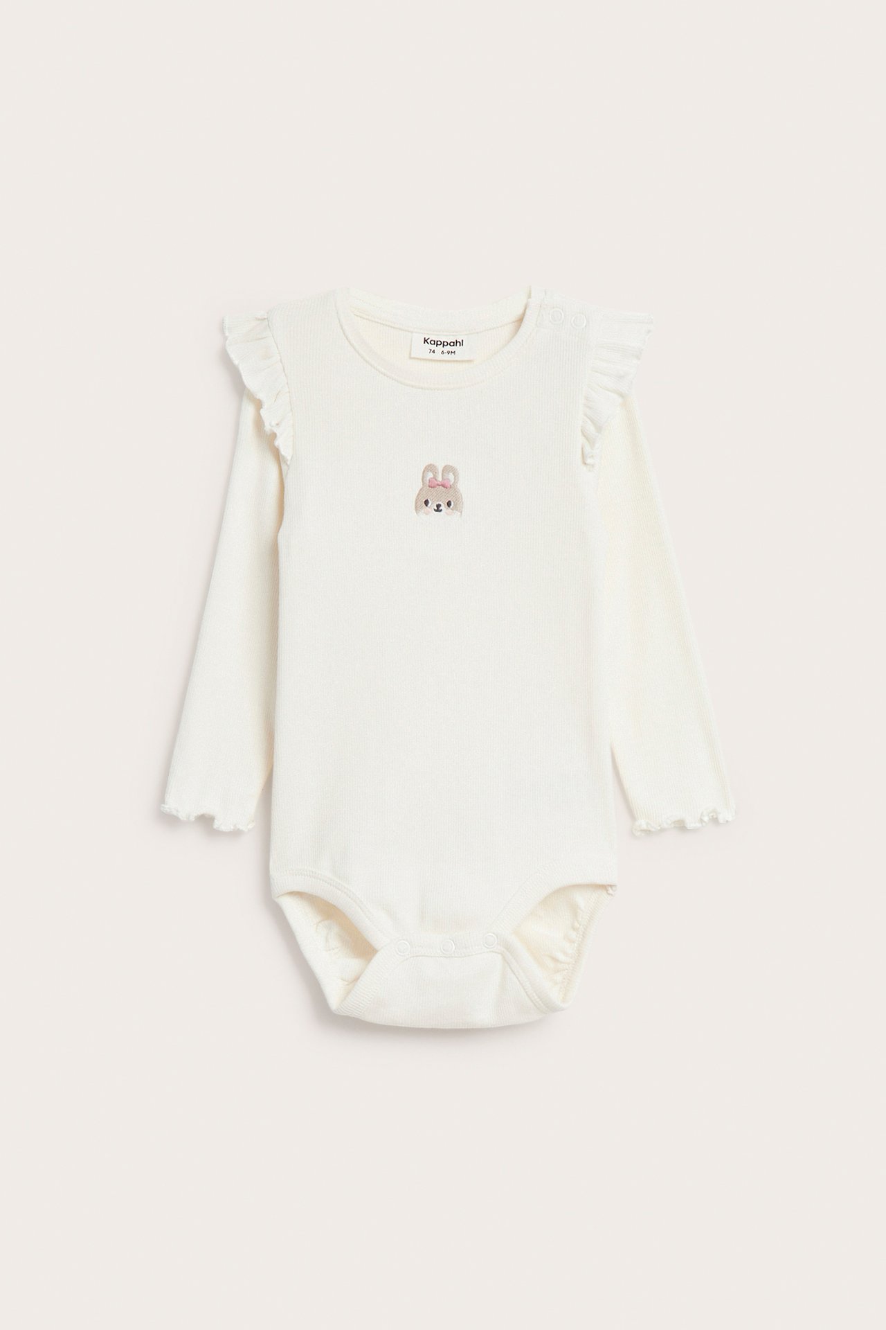 Framsida av en benvit långärmad babybody med axelvolanger och en broderad kanin.