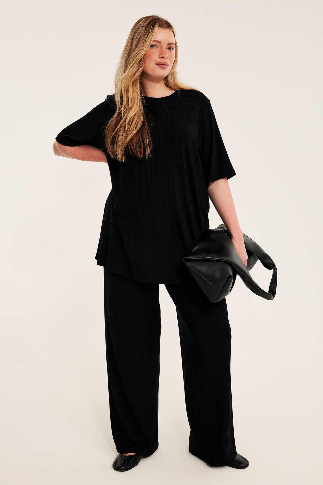 Framsida: Kvinna i svart ribbad oversized t-shirt och vida byxor, håller en svart väska.
