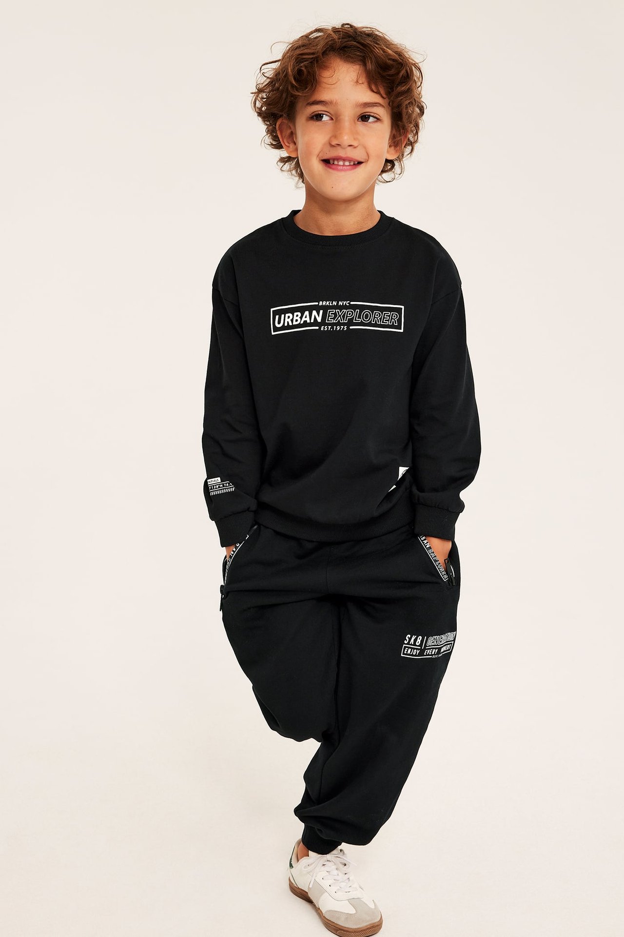 Frontvy av ett barn i svart bomullssweatshirt med "Urban Explorer"-tryck, svarta mjukisbyxor och vita sneakers.