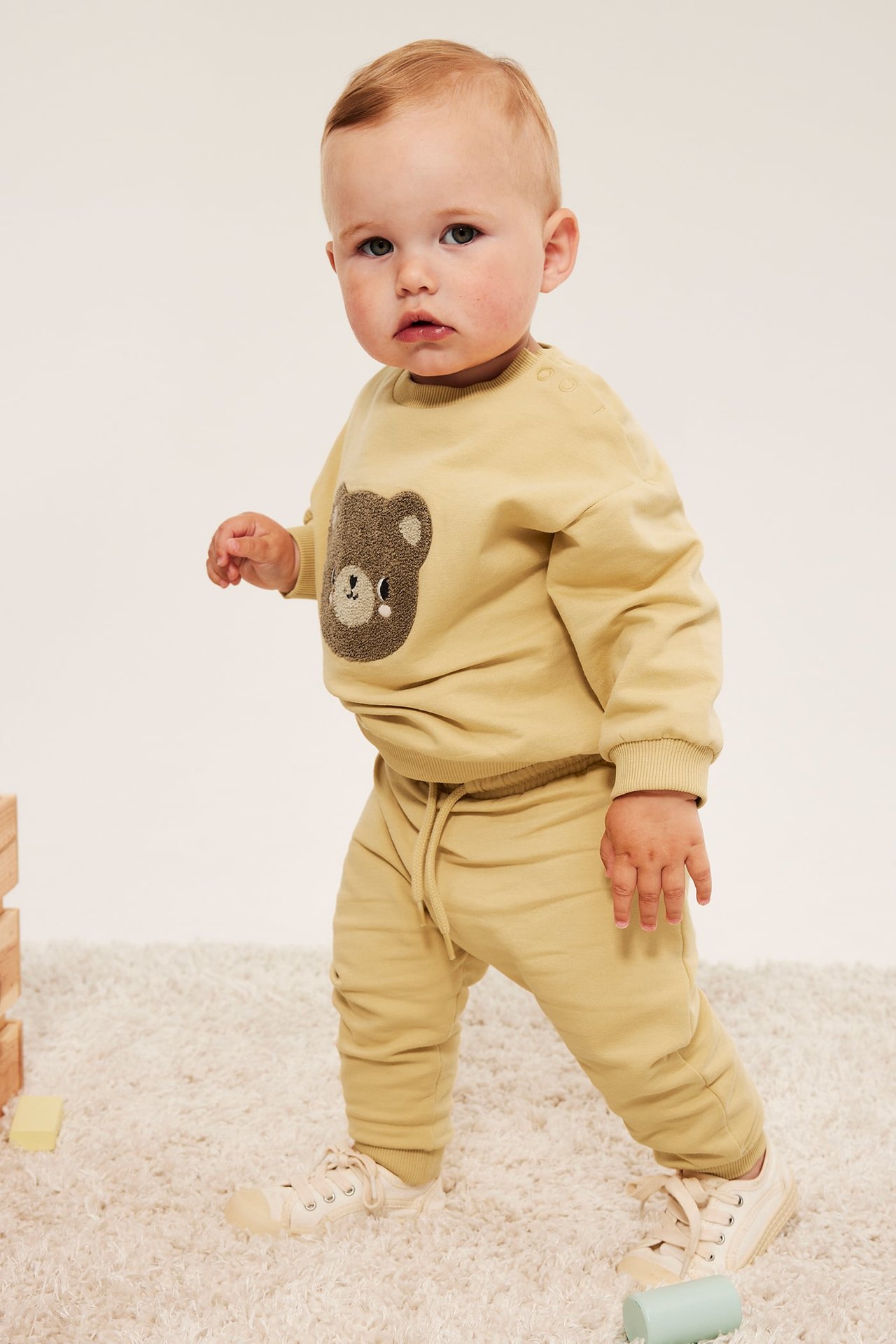 Forfra: baby har lys gul genser med bjørn, matchende joggebukse og lyse joggesko.