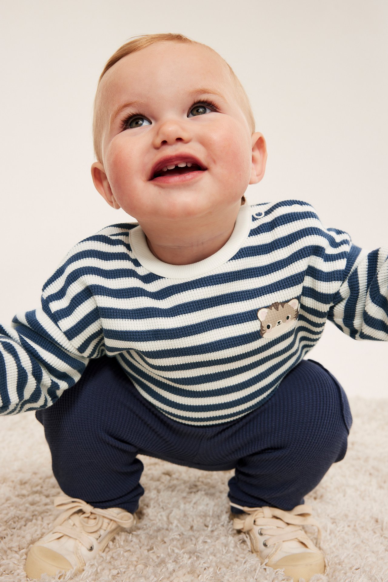 Framsida på baby med blåvitrandig våfflad tröja, marinblå byxor och beiga sneakers.