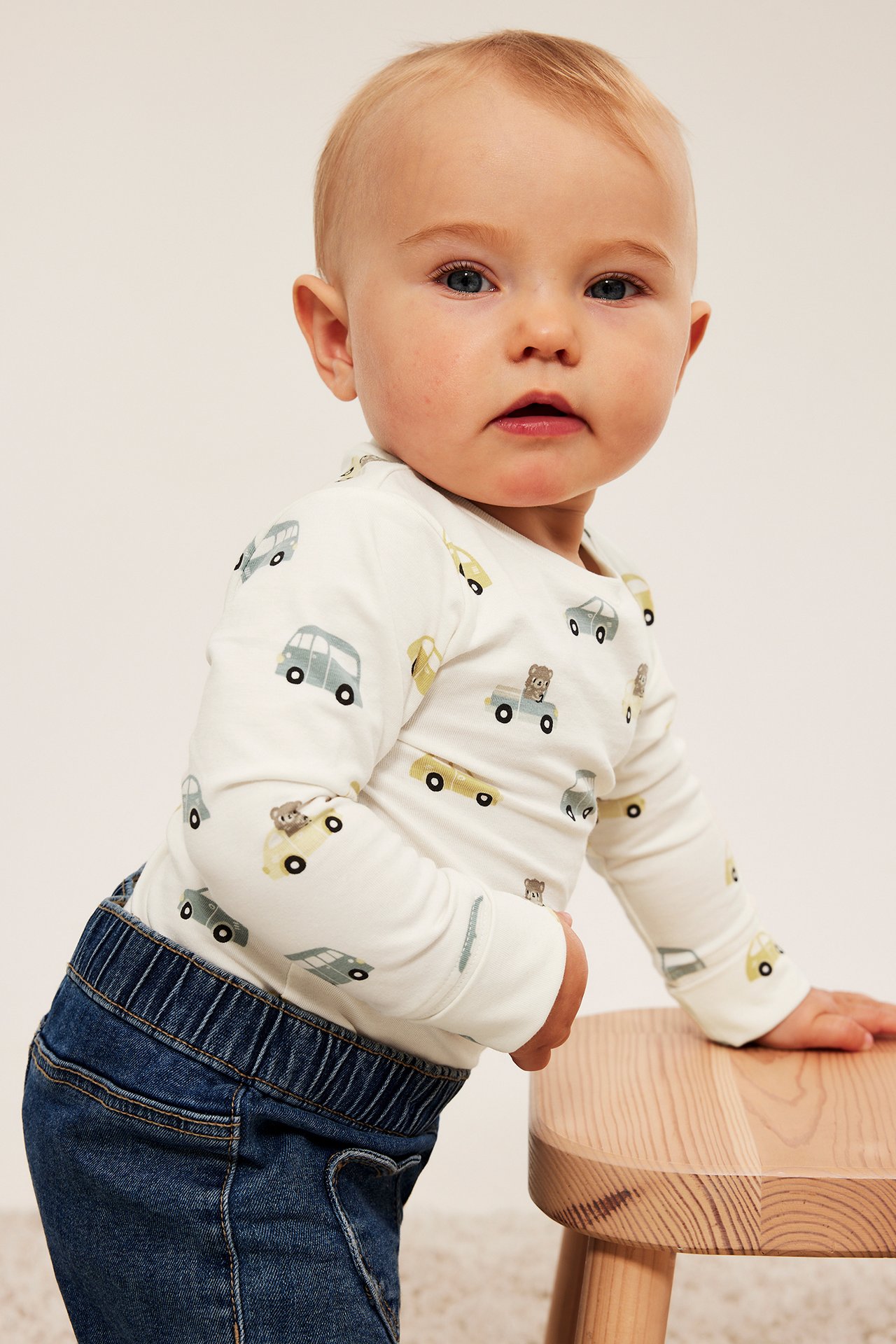 Front: baby i hvit langermet body med bilprint og blå jeans.