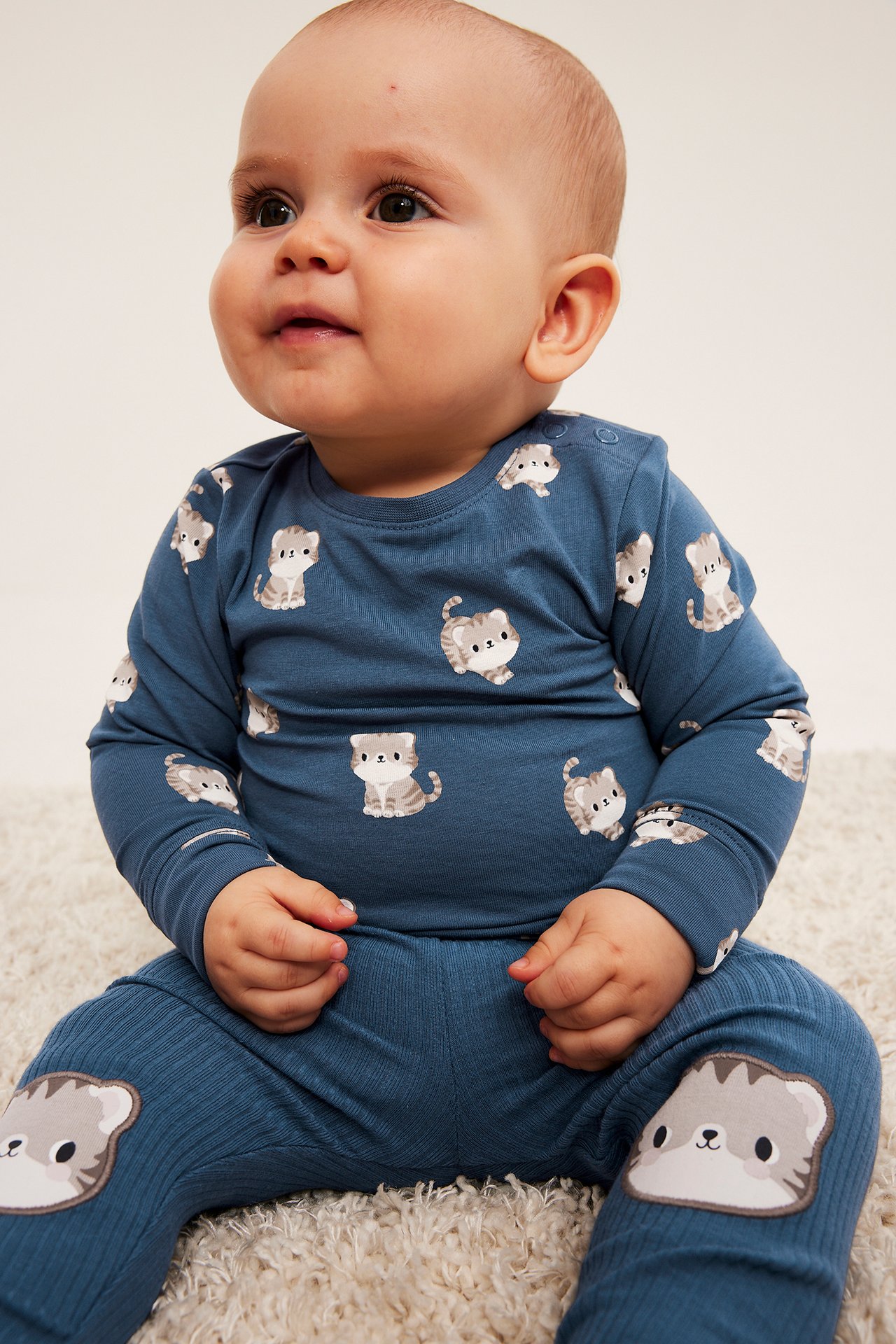 Framifrån: baby i blå långärmad tröja och ribbade byxor med kattmönster.
