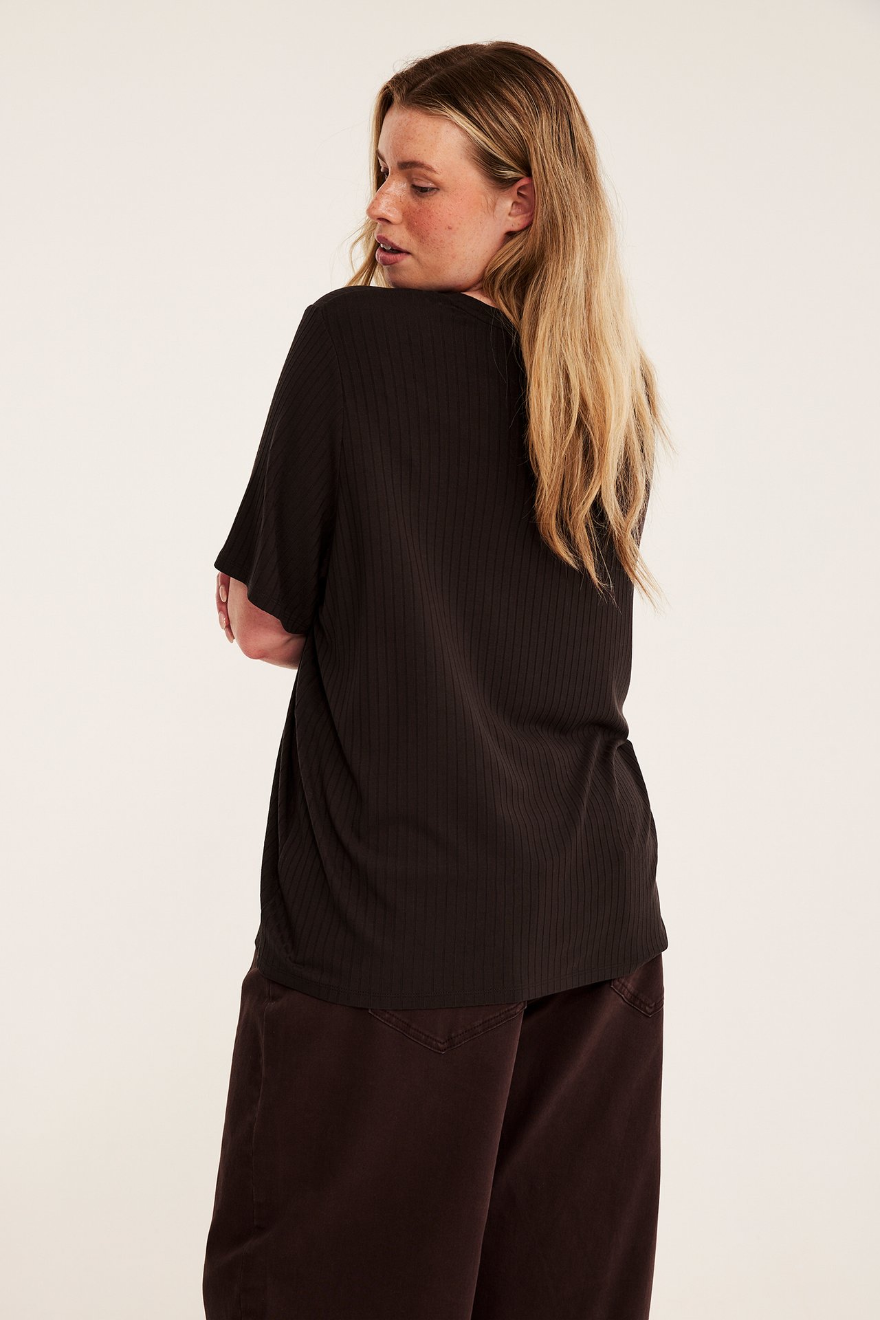 Bakre/sidovy av en kvinnlig modell i svart ribbad kortärmad topp och mörkbruna vida byxor.