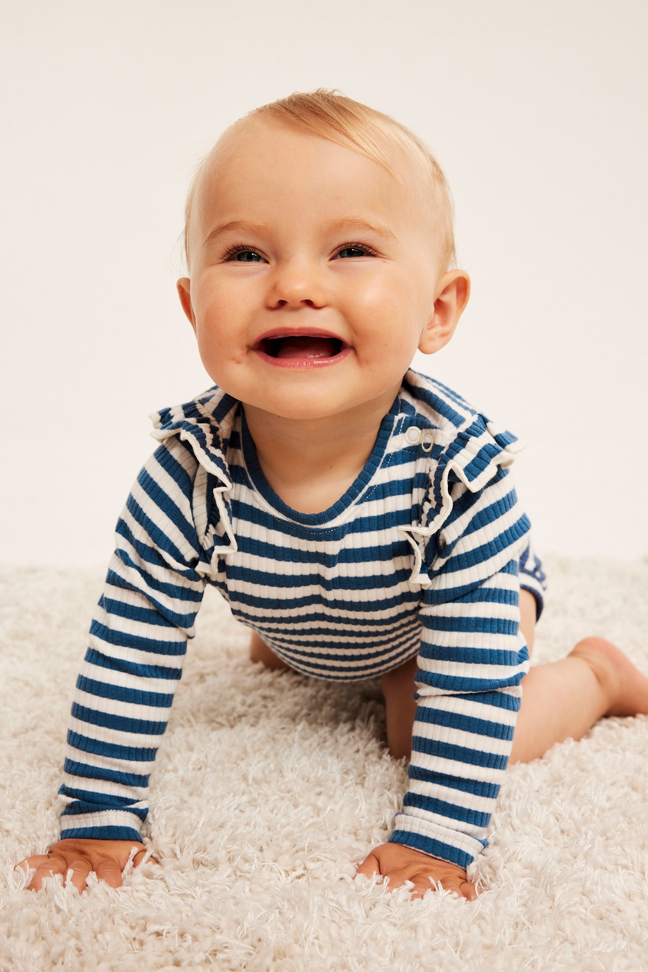 Forfra, baby kryper i blå og hvitstripet langermet ribbet body med skulderkapper.