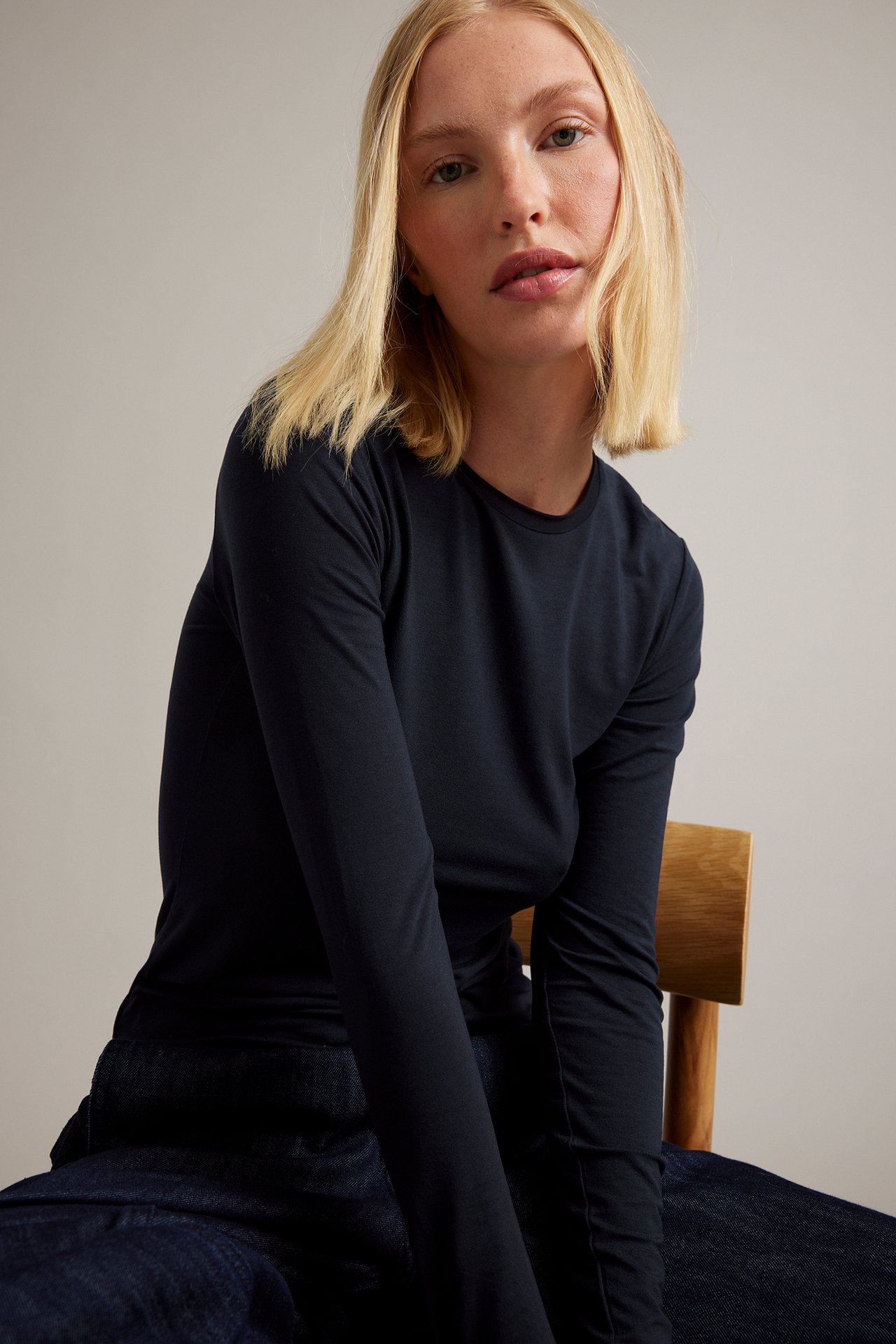 Frontbilde av sittende kvinne i marineblå langermet topp med rund hals og mørkblå jeans.