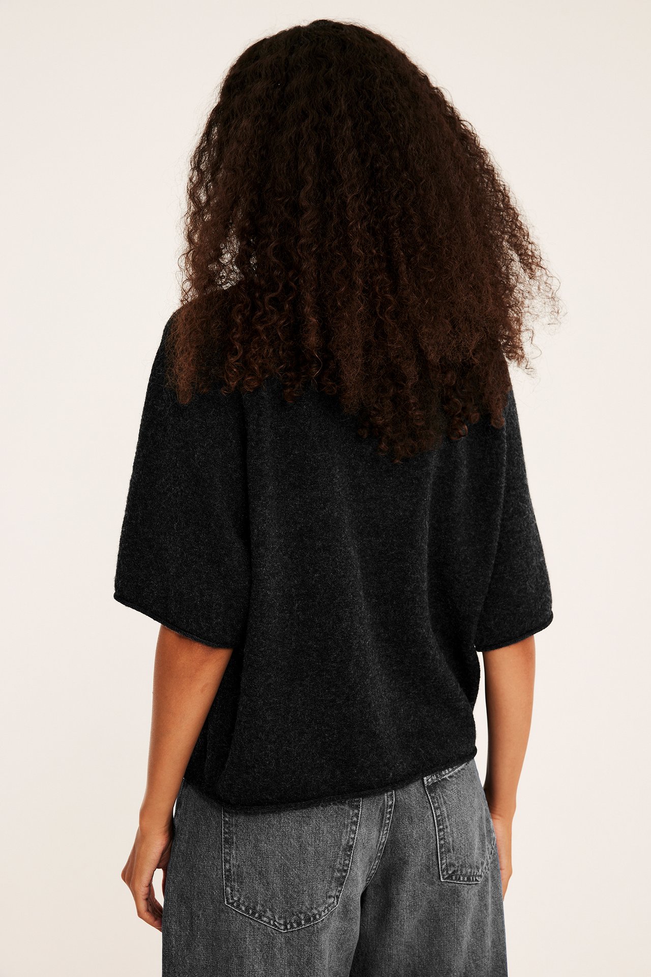 Ryggvisning av kvinne iført mørkegrå kortermet strikkegenser og mørkegrå jeans.