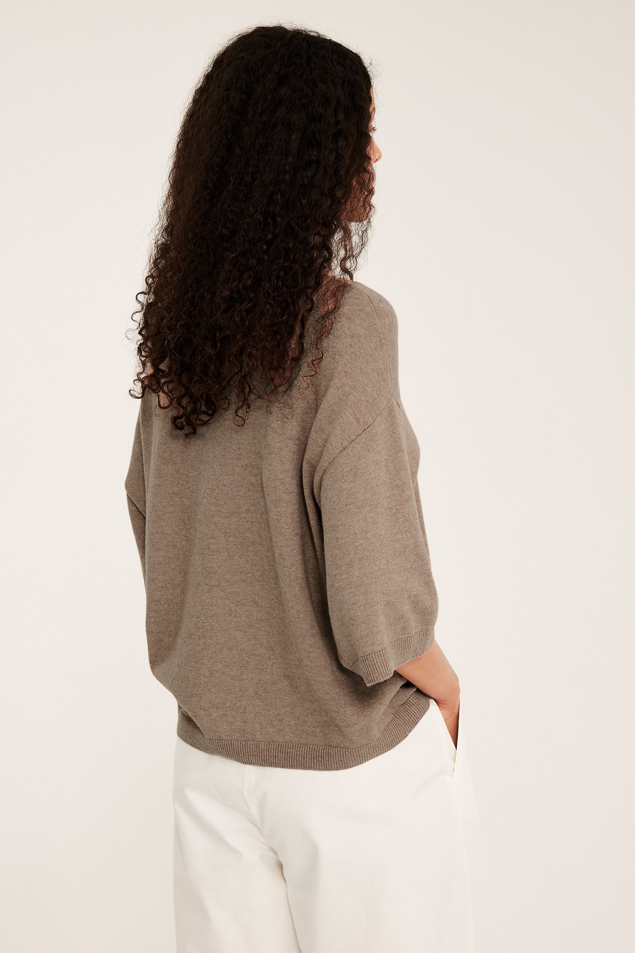 Nainen takaapäin yllään beige meleerattu oversized lyhythihainen neuletoppi ja valkoiset housut.
