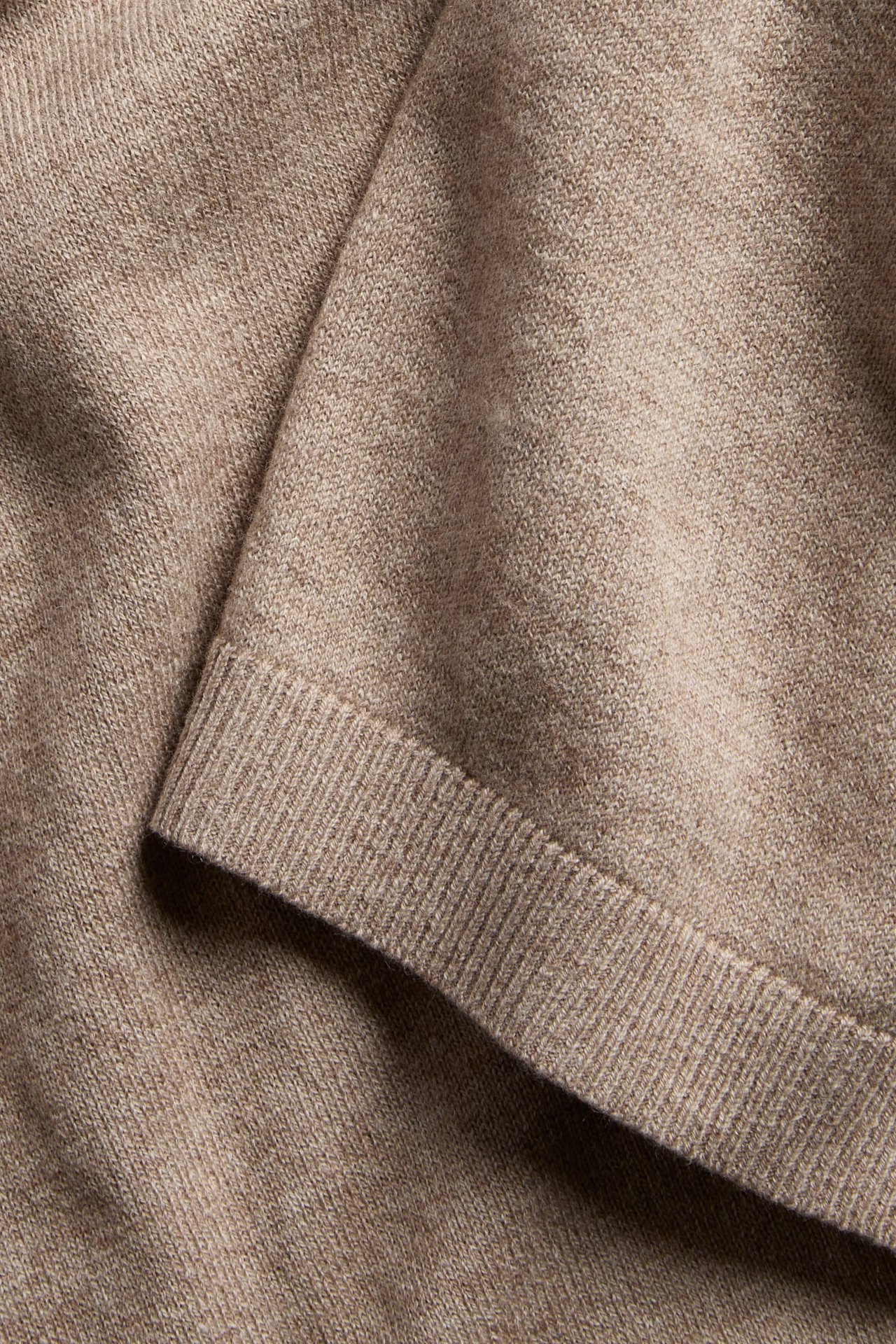 Zbliżenie na beżowy melanżowy sweter z mieszanki wełny z widocznym ściągaczem u dołu.