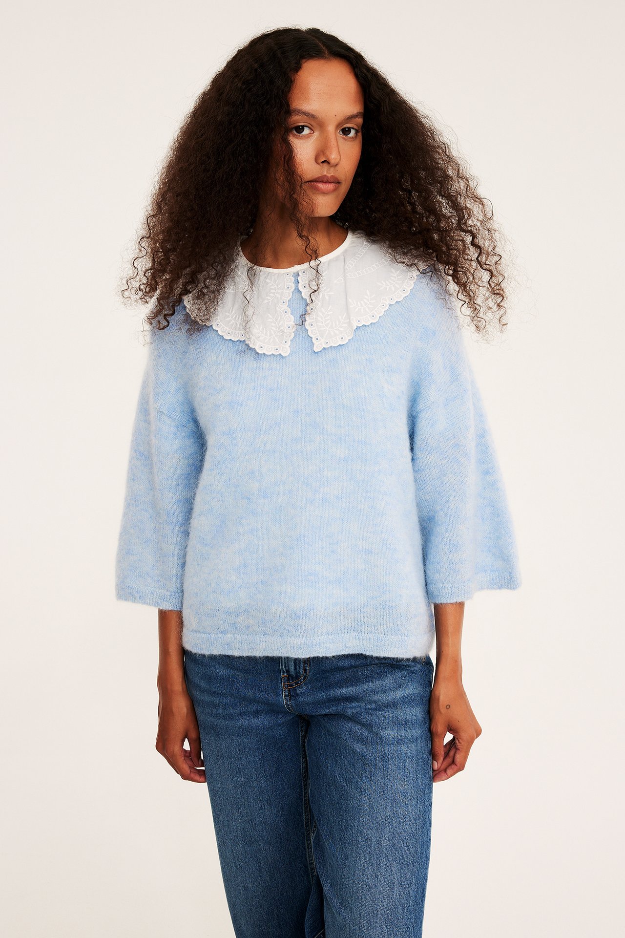 Forfra, kvinnelig modell i lyseblå lodden genser med brodert hvit krage og blå jeans.