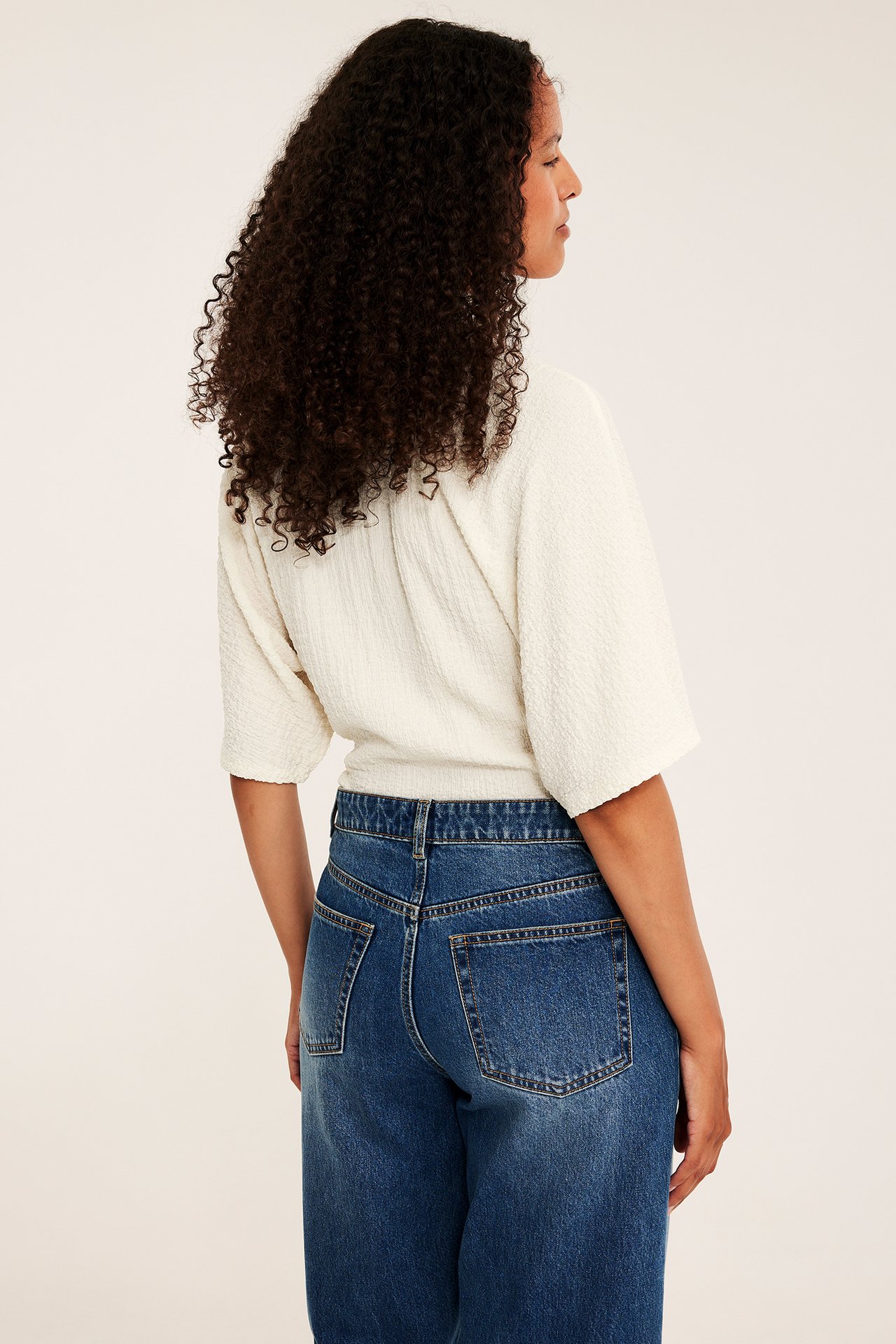 Baksida av kvinna i krämvit kortärmad strukturerad omlott-topp och blå jeans.