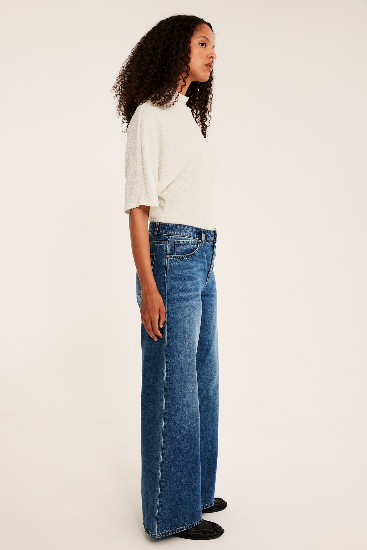 Sidovy av kvinnlig modell i vida blå jeans, krämvit texturerad topp och svarta ballerinaskor.