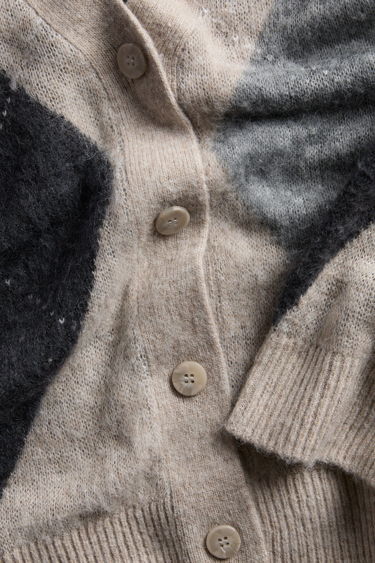 Nærbilde av en beige, svart og grå mønstret strikket cardigan med lyse knapper.