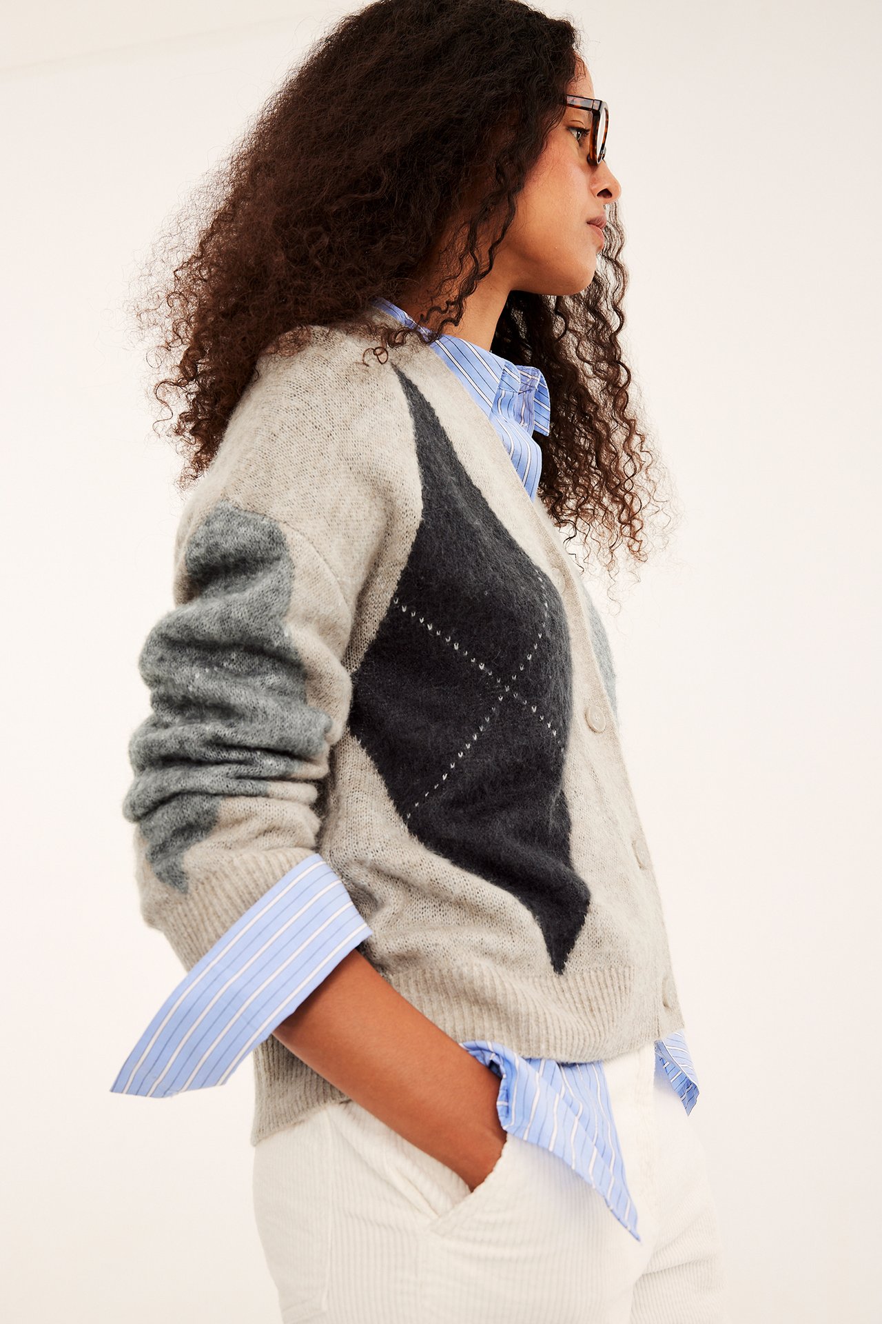 Sidebilde av kvinne i beige argyle strikket cardigan, blåstripet skjorte og hvite bukser.