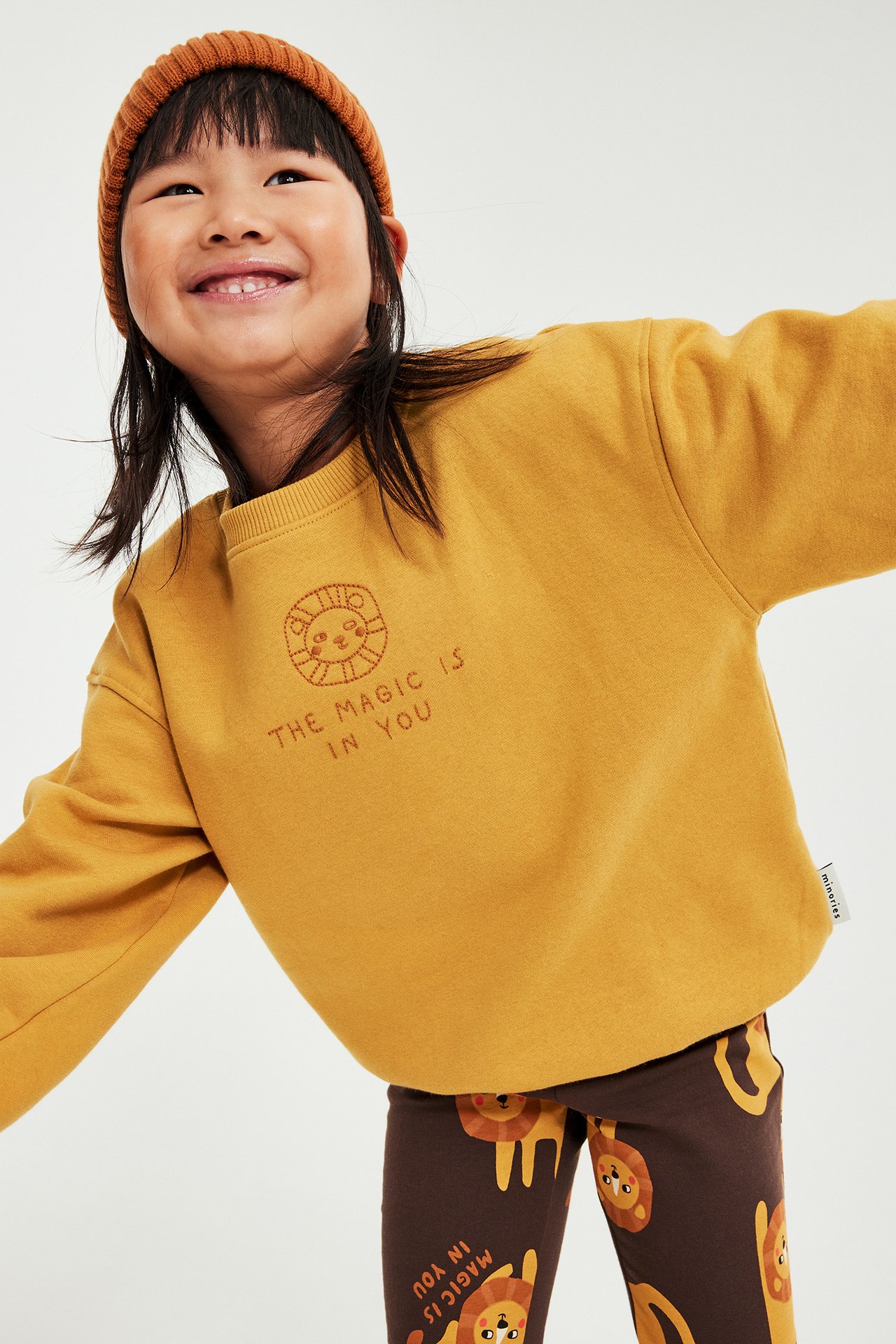 Glad unge i senapsgul sweatshirt med lejontext, bruna leggings med lejontryck och rostfärgad mössa.