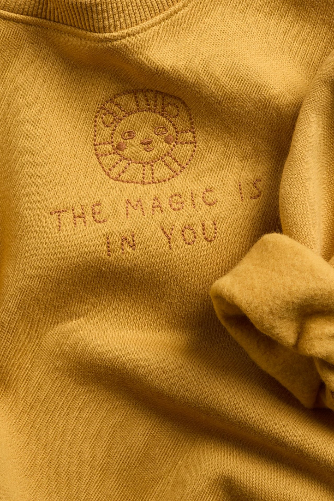 Närbild av en senapsgul sweatshirt med ett broderat solansikte och texten "THE MAGIC IS IN YOU".
