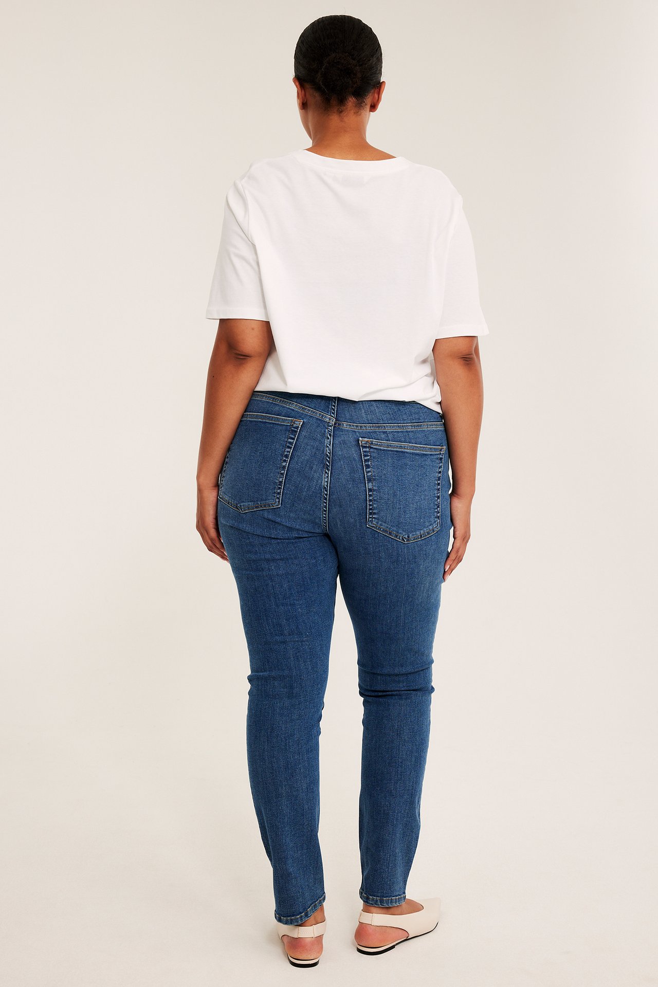 Takaapäin näkymä naisesta sinisissä slim-fit farkuissa, valkoisessa t-paidassa ja beigissä kengissä.