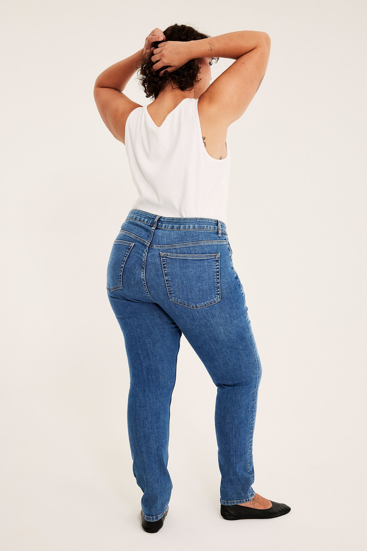 Ryggbilde av kvinne i blå rette jeans, en hvit singlet og svarte ballerinasko.