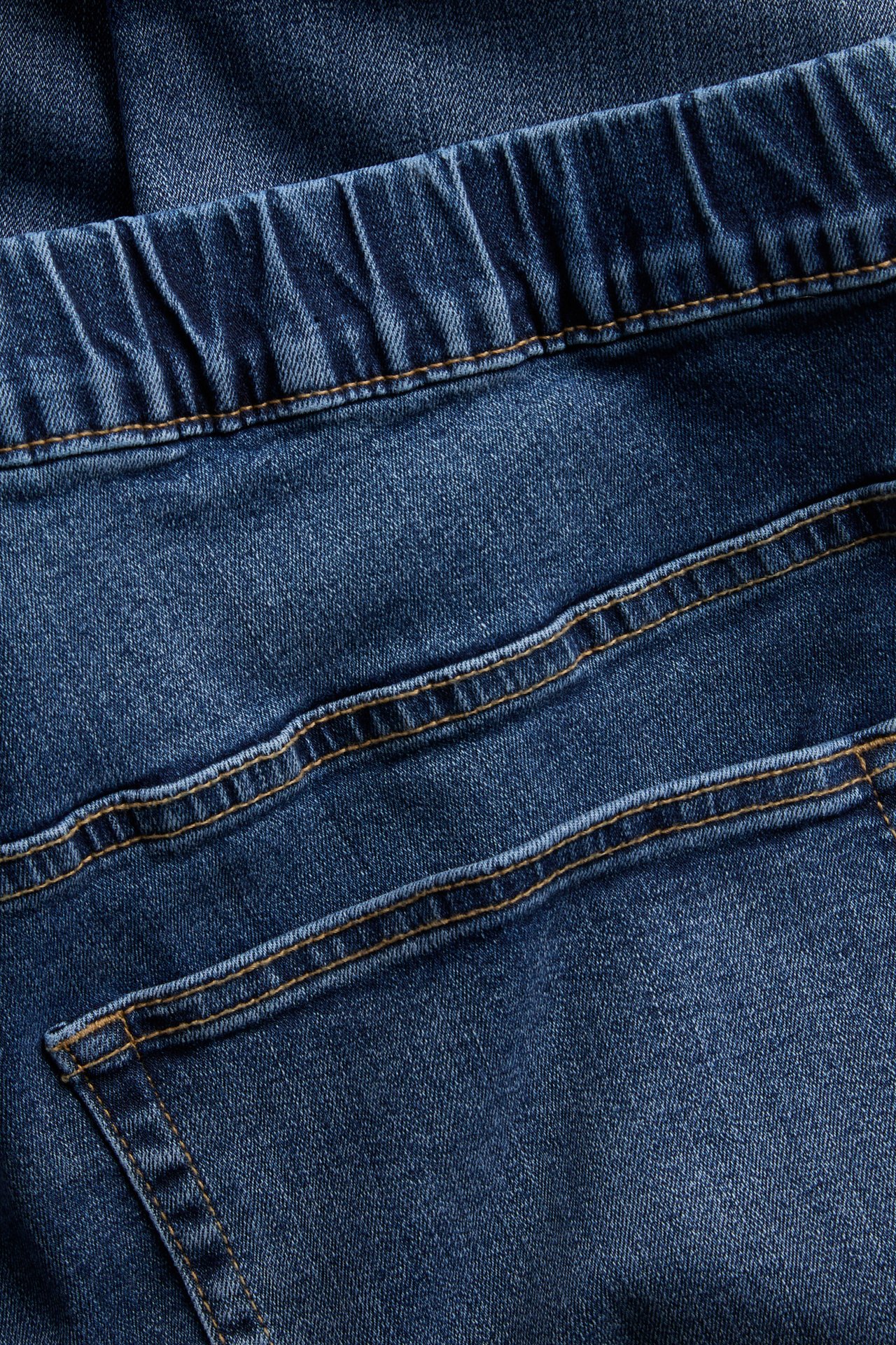 Nærbilde av blå denimjeans med elastisk midje, kontrastsømmer og baklomme.