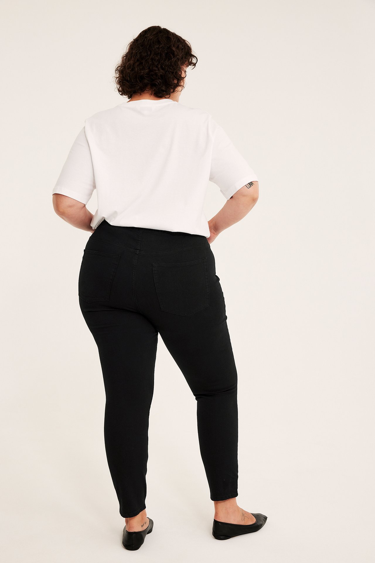 Kvinne i svarte skinny jeans, en hvit t-skjorte og svarte flate sko, bakfra.