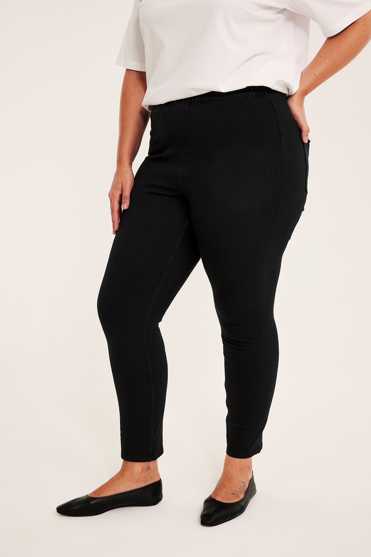 Sidebilde av en kvinne iført svarte pull-on skinny jeans, en hvit topp og svarte ballerinasko.