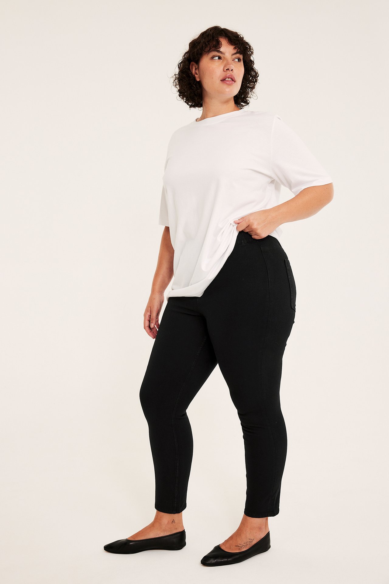 Sidebilde av plus-size kvinne i svarte skinny jeans, hvit t-skjorte, svarte ballerinasko.
