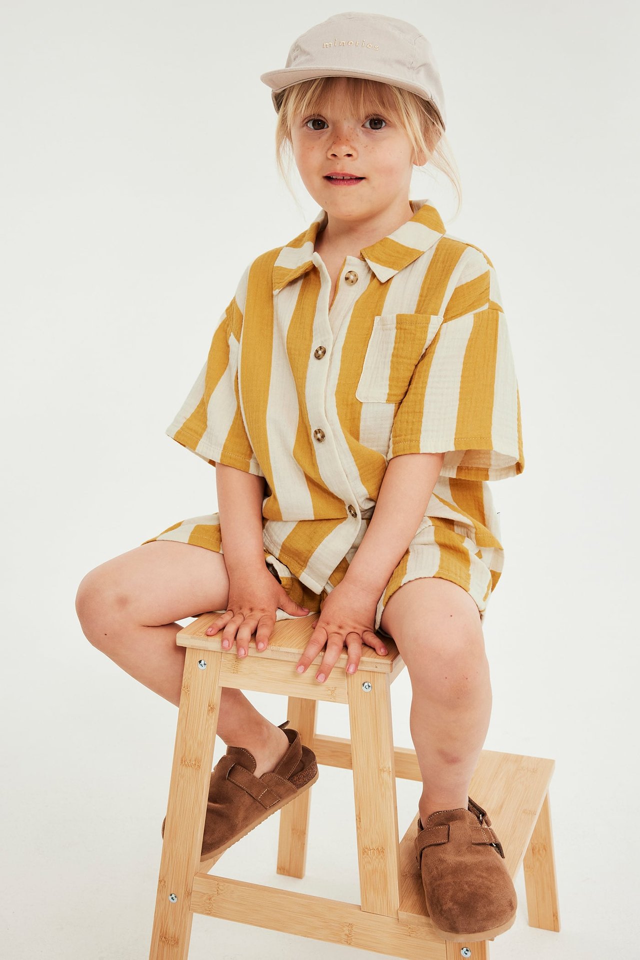 Frontbilde: barn i stripet gult og hvitt musselinsett, beige kaps og brune sko, sitter på krakk.