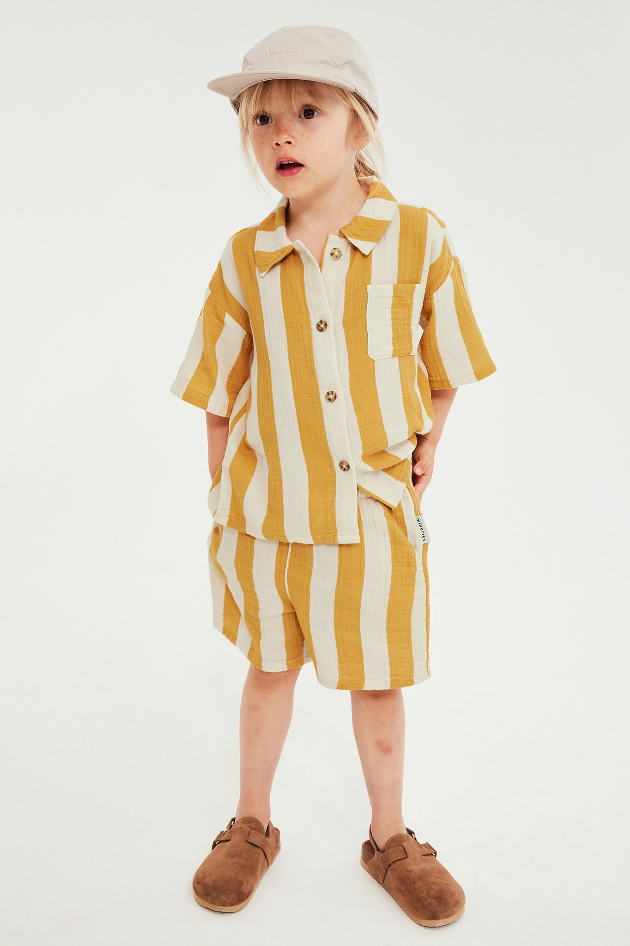 Front view: kid i randigt set av gul och vit muslin, beige keps och bruna tofflor.