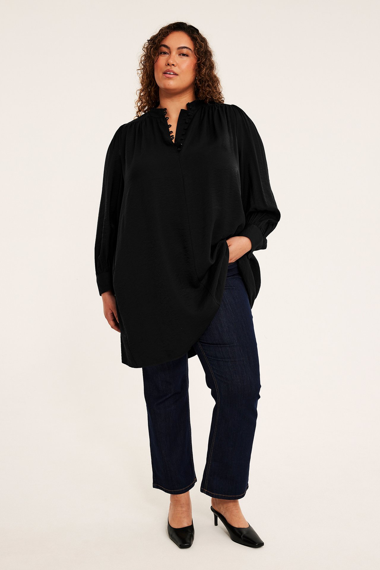 Framifrån: kvinnlig modell i svart satintunika, mörkblå jeans och svarta muleskor.