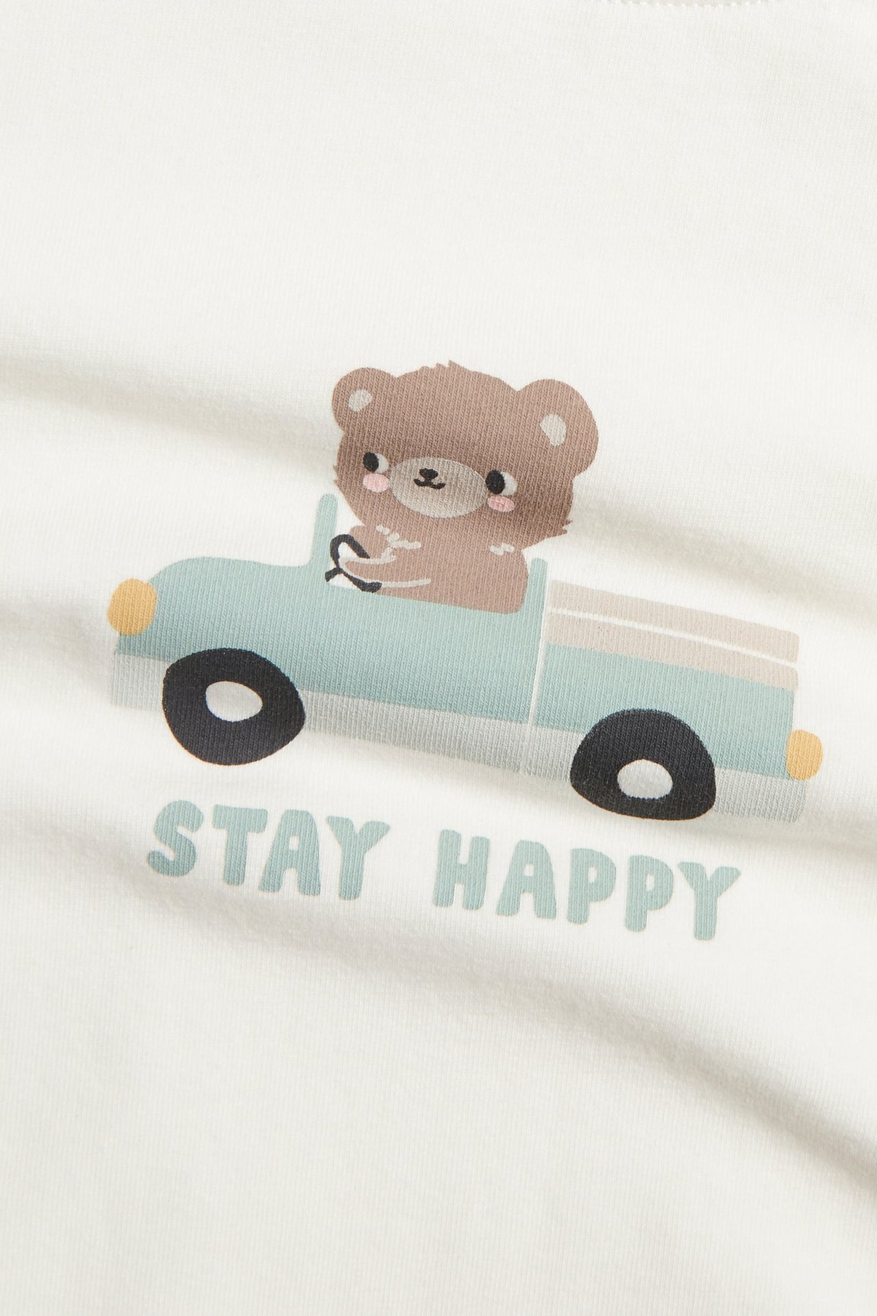 Nærbilde av hvit topp med trykk av bjørn i blå lastebil og 'STAY HAPPY' tekst.