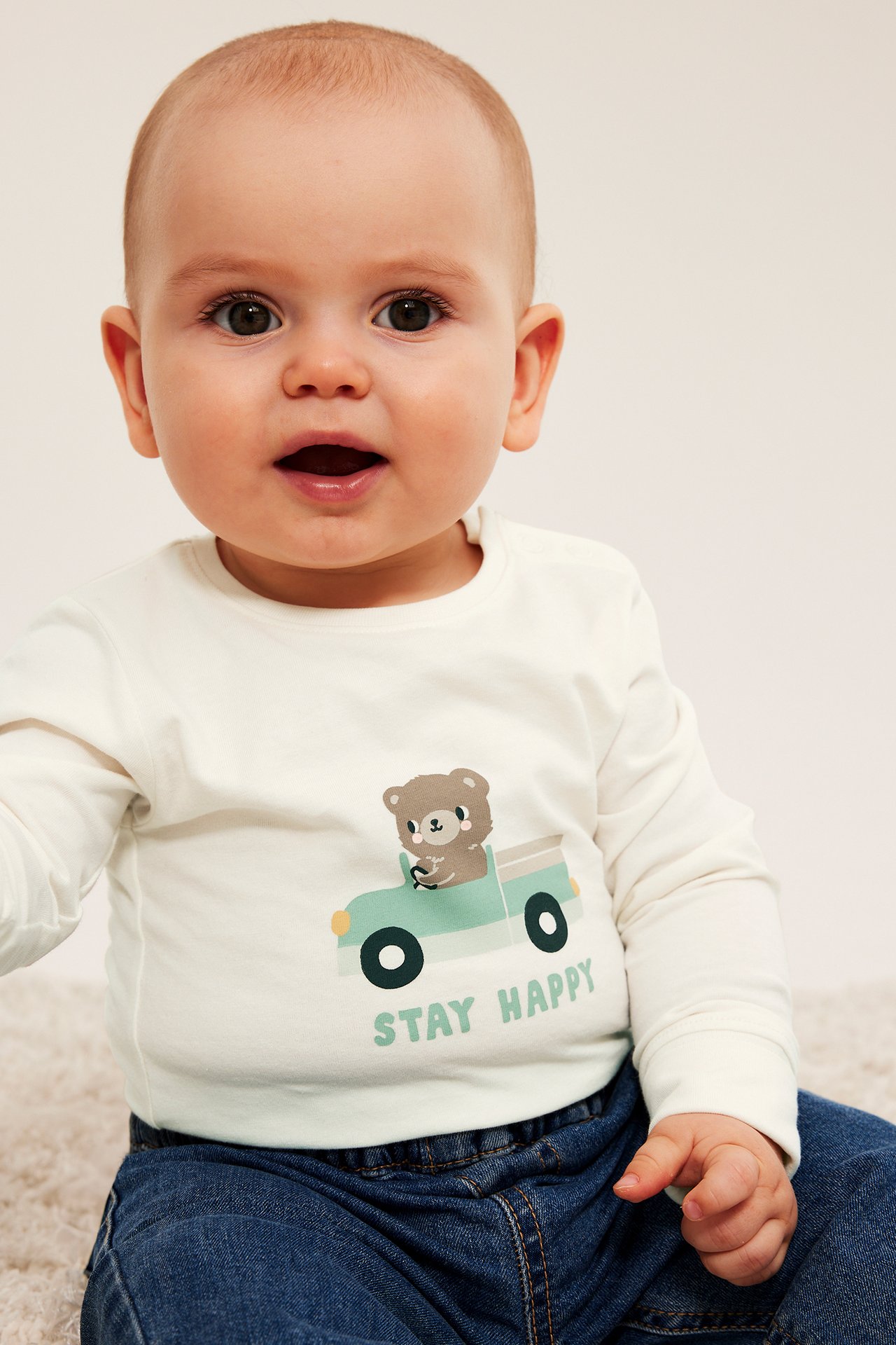 Framifrån en baby i en gräddvit långärmad tröja med björnmotiv och mörkblå jeans.