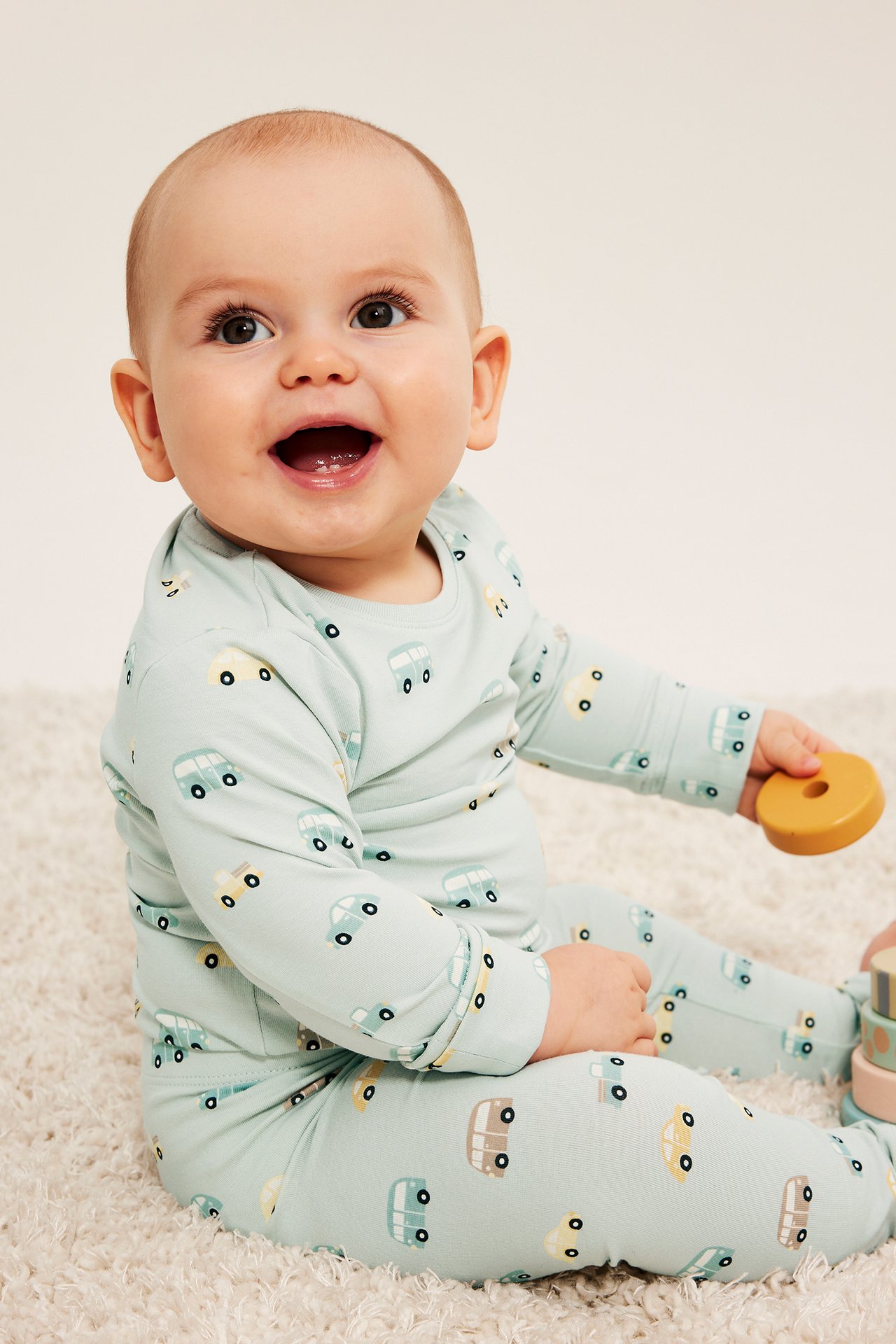Framifrån: baby i ljusblå långärmad pyjamas med bilmönster, håller en träring.