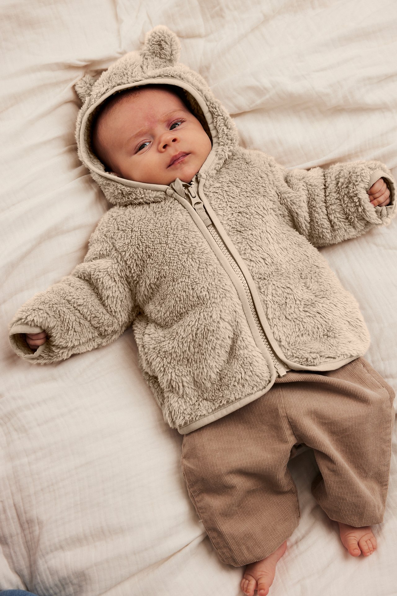 Framsida på bebis i beige fluffig huvjacka med öron och bruna manchesterbyxor.